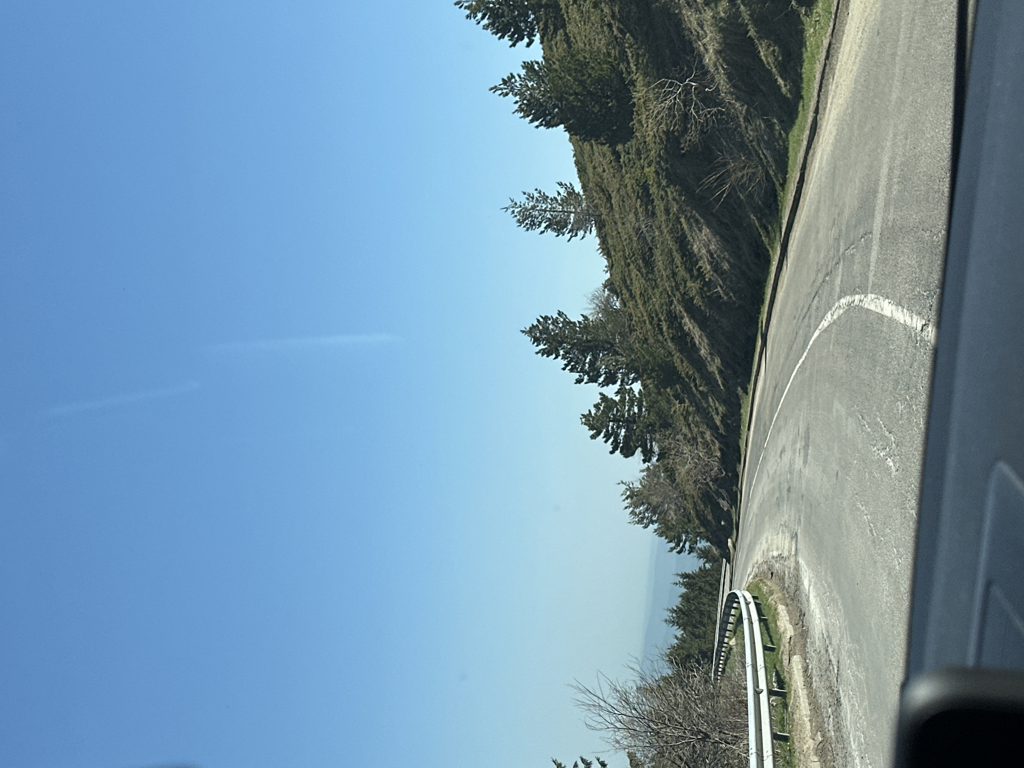 A road curves uphill through a wooded area near Beli Osam, Bulgaria. A metal guardrail flanks the edge of the road on the right. The sky is clear.
