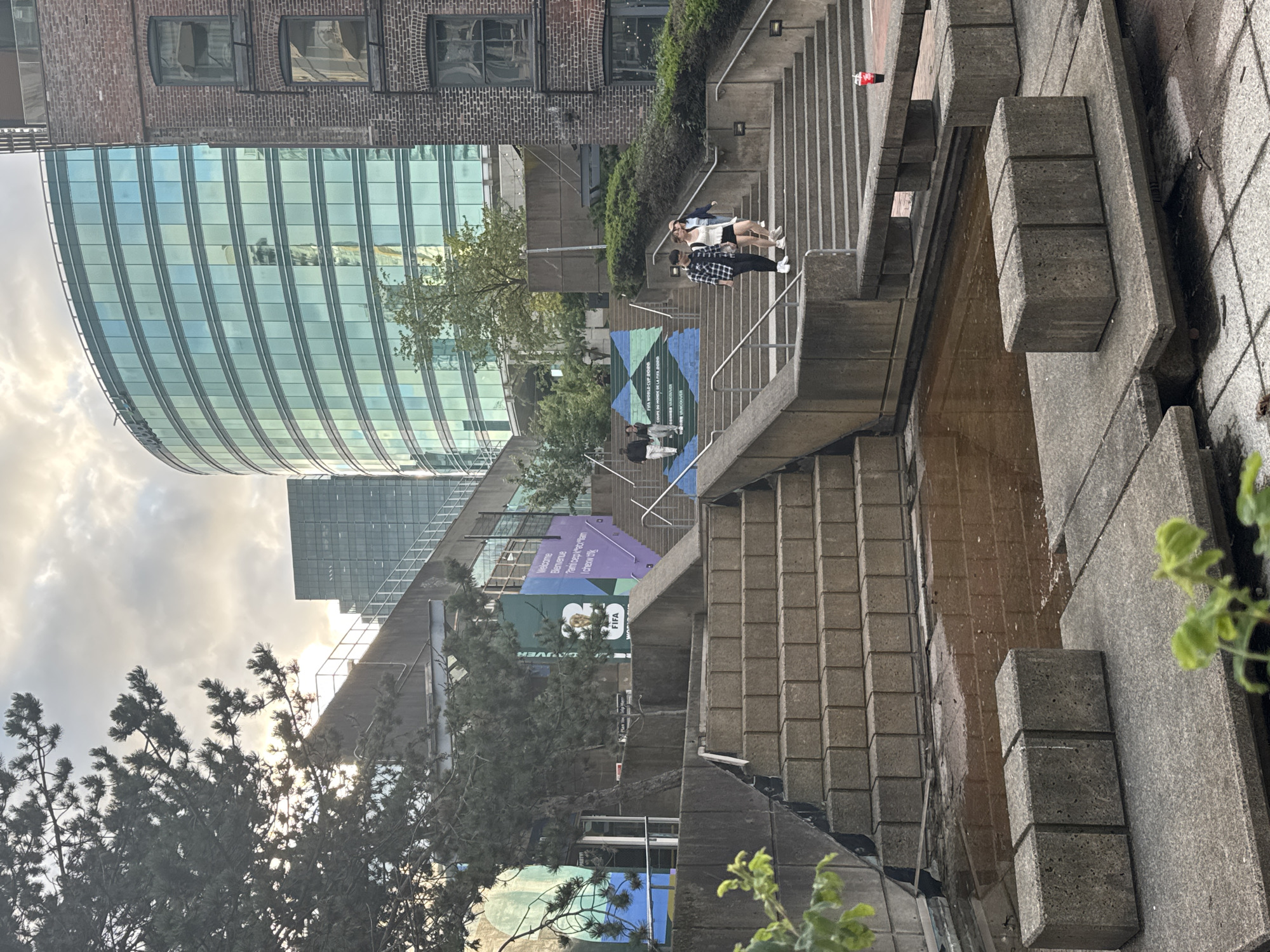 The image shows an outdoor plaza with concrete structures and stairs in Vancouver, Canada. A large, curved, glass-facaded building is visible in the background, reflecting the sky and surrounding structures. Adjacent to it is a brick building with visible windows. On the stairs, there are a few individuals walking, with some people interacting. In the foreground, there is a shallow pool of water surrounded by concrete blocks and planters with foliage. A banner on the stairs displays "FIFA WORLD CUP 2026" and "VANCOUVER". The lighting suggests it is late afternoon or early evening with the sun low in the sky.