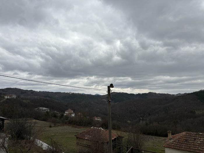 The image depicts a wide panoramic view of a rural, hilly landscape in Bukovo, Bulgaria, under a heavily overcast sky. The sky is uniformly gray with dense cloud cover. In the foreground, a prominent utility pole is positioned left of center, supporting two dark horizontal power lines that traverse the mid-ground. Bare deciduous trees and dormant shrubs with visible branches are present in the immediate foreground, alongside sections of a light-colored structure or wall on the lower left. To the far right in the foreground, a portion of a building with a reddish-brown tiled roof and a light-colored wall is visible. The mid-ground features dry, yellowish-green grassy patches interspersed with dark, leafless trees covering rolling hills. Several residential structures with light-colored facades and reddish-brown tiled roofs are scattered across the hill slopes. The background consists of successive layers of wooded hills, appearing dark due to the tree cover and the overcast conditions, receding into the distance. No individuals or active subjects are discernible within the frame.