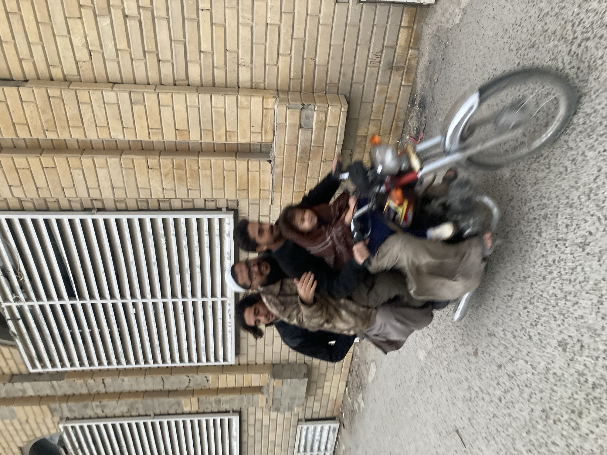 A low-angle medium shot shows four people riding a motorcycle on a street in Quetta City Tehsil, Pakistan. The motorcycle is moving, as suggested by the blurred background and the rotation of the front wheel. A girl in a brown leather jacket sits behind the driver, who wears a black jacket. Behind them, another man wearing a camouflage jacket sits on the back of the motorcycle. The building on the left features window grills and tan bricks.