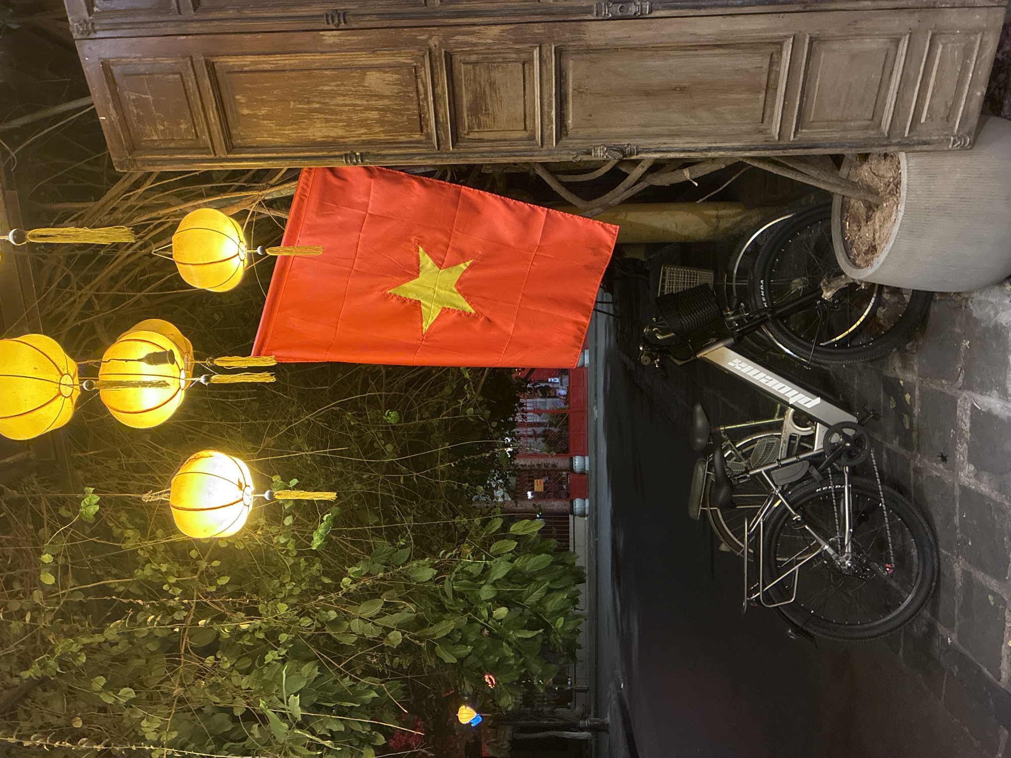 The image captures a night scene in the city of Hội An Ward, Vietnam. Dominating the view is a vibrant red Vietnamese flag with a yellow star, positioned against the backdrop of an ornate wooden cabinet or structure. Strung lanterns emit a soft yellow light from trees, casting a warm glow. Below, a bicycle is parked on a brick-paved surface, and a potted plant sits nearby. The red facade of a building is visible in the distance.