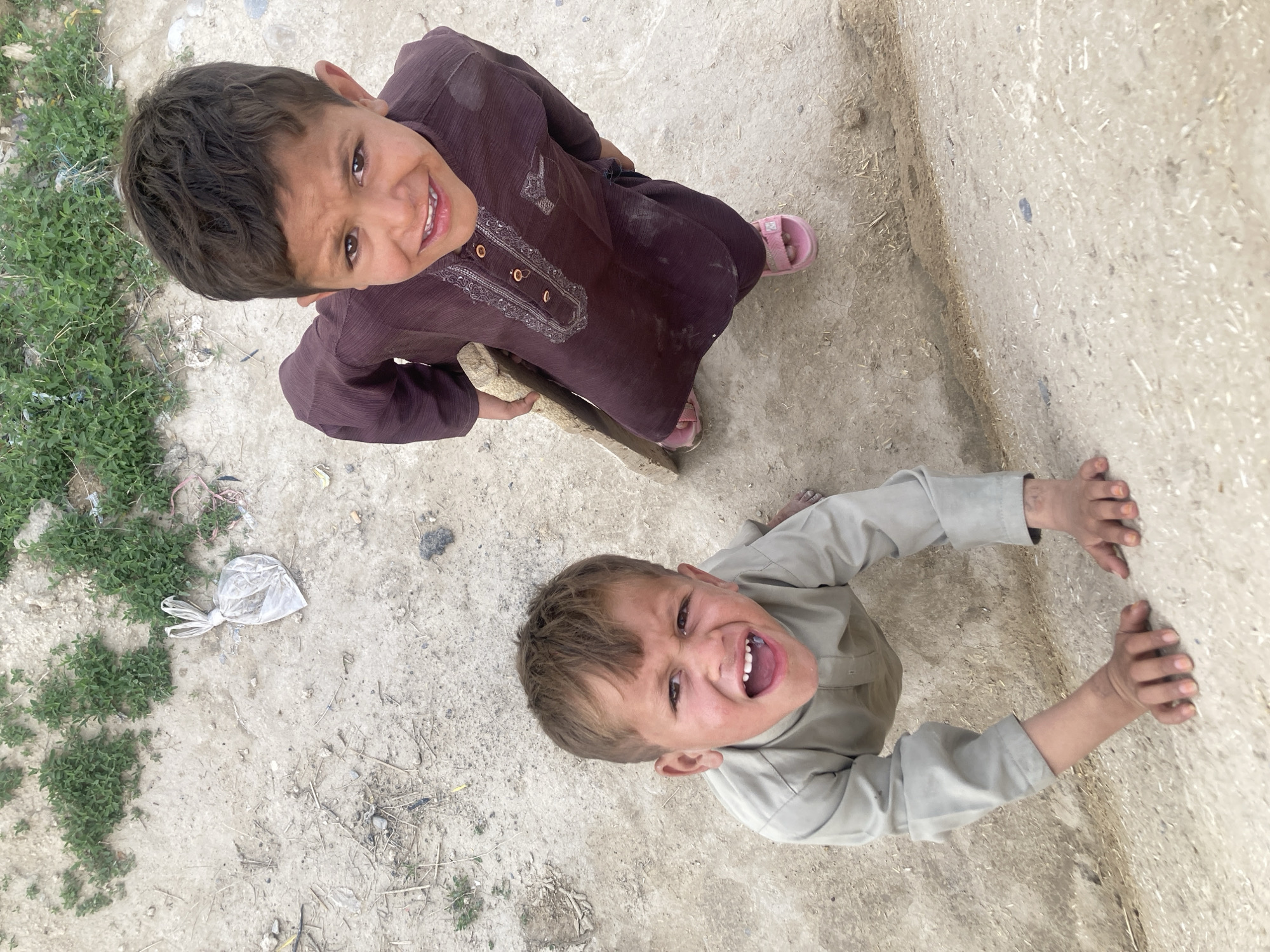 The image captures two young male children outdoors on dirt ground in Quetta City Tehsil, Pakistan. The photograph is taken from a high-angle perspective, looking down at the subjects. On the left, a child approximately 3-5 years old wears a light olive-tan full-sleeved tunic (shalwar kameez top). He is barefoot, with both hands placed on a light-colored, raised concrete or dirt edge in the immediate foreground. His mouth is open wide, revealing teeth, and his eyes are wide, indicating an expressive vocalization or strong emotion. On the right, a child approximately 5-7 years old wears a dark reddish-brown full-sleeved tunic with embroidered detailing on the placket and cuffs. He wears pink open-toed sandals and is seen holding a rough, light-colored wooden plank with his left hand. His expression is a faint smile directed towards the camera. Both children have visible dirt on their faces and clothing. The environment consists of dry, light brown soil with scattered small pebbles, dried plant fragments, and minor debris. In the upper left background, a crumpled, clear plastic bag, tied at the top, lies on the ground, surrounded by patches of green, low-growing vegetation. The scene is illuminated by natural daylight.