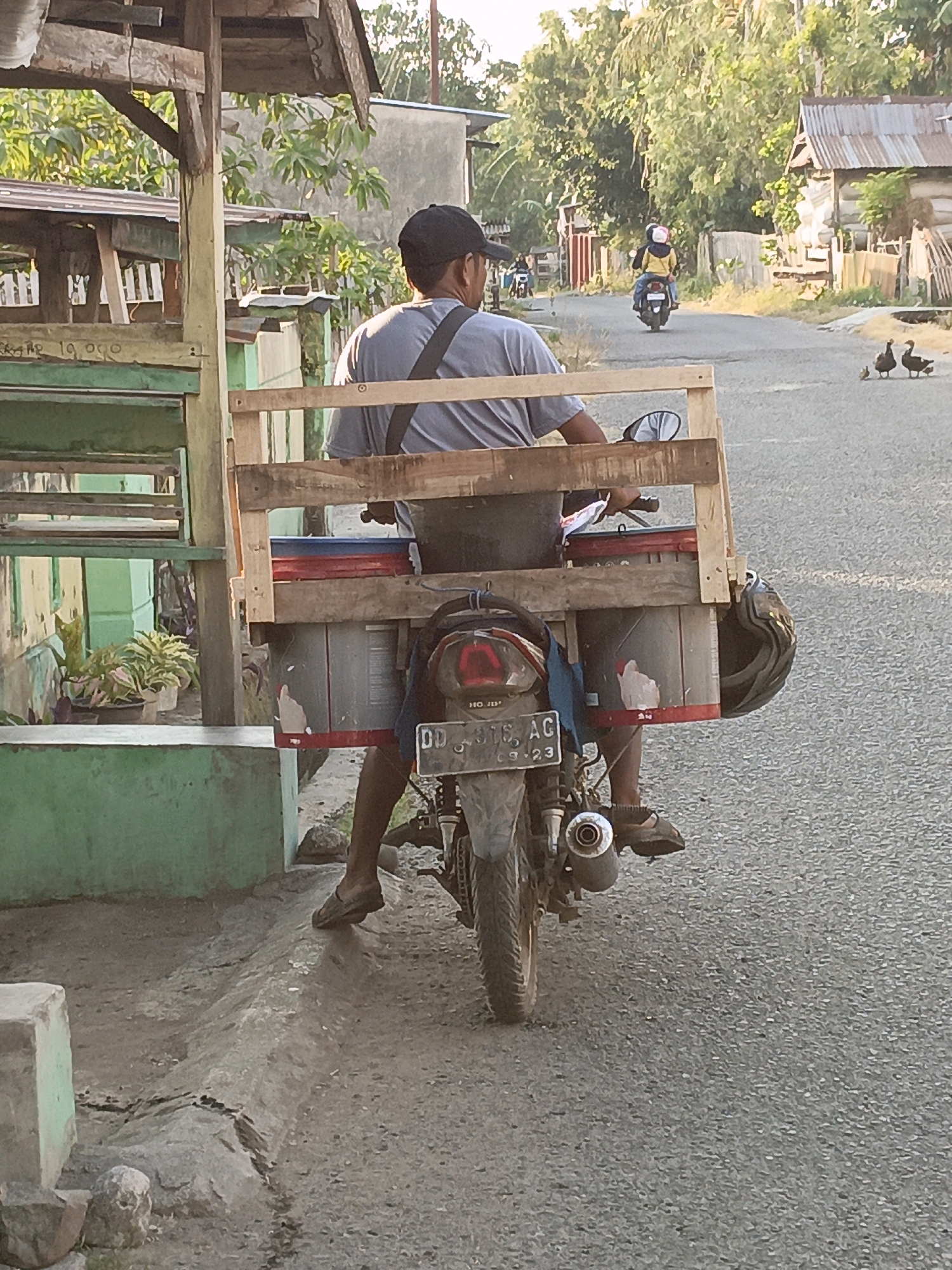 In Bulukumba, Indonesia, a man wearing a dark baseball cap and a grey t-shirt is seated on a motorcycle, viewed from behind. He is carrying a wooden structure on the back of the motorcycle, composed of upright planks and horizontal supports, which holds two large cylindrical containers. A dark helmet is attached to the right side of this structure. The motorcycle has a license plate that reads "DD 3918 AC" and "09-23". The man is wearing dark shorts and dark shoes. In the background, another motorcycle with two riders is visible on the asphalt road, and further in the distance, there are buildings with corrugated metal roofs and trees. A group of ducks, one adult and two ducklings, are present on the right side of the road. The scene appears to be a rural or semi-urban street during daylight.