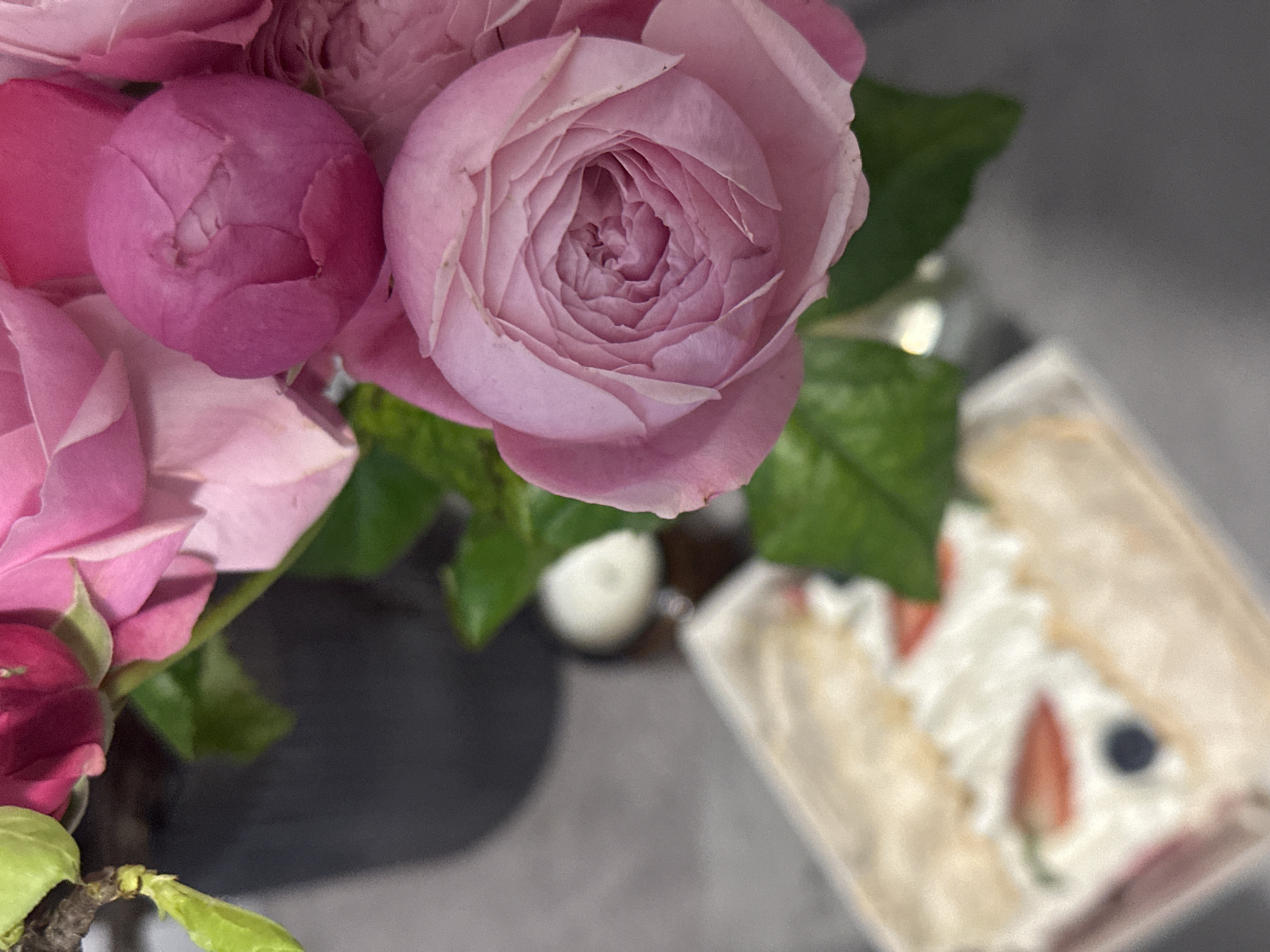 A high-angle shot displays a cluster of pink roses with green leaves, positioned to the left and partially obscuring the view of a boxed cake. The cake, decorated with white cream and sliced strawberries, has a single visible blueberry. The overall color palette is soft, dominated by pinks, greens, and white. The backdrop appears to be a gray surface. The image does not provide enough information to determine the location as Kazan, Russia.