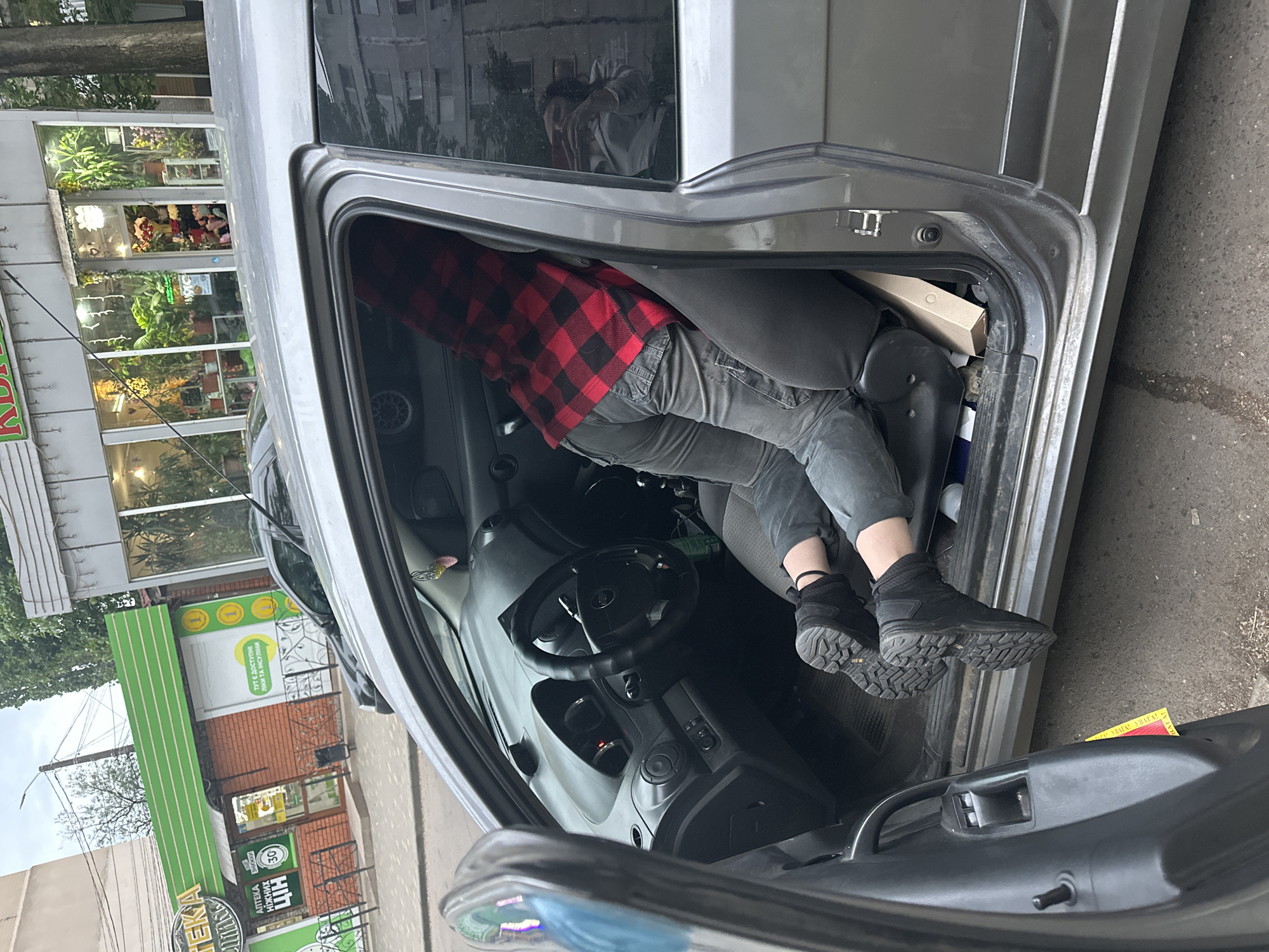 The image captures a person partially inside a grey car, parked on what appears to be a street or sidewalk next to commercial buildings.
The main subject is a person, visible from the torso downwards, leaning into the driver's side of the car. They are wearing a red and black plaid shirt, grey jeans, and sturdy black ankle boots with thick soles. Their posture suggests they are either reaching for something inside the vehicle or in the process of entering/exiting.
The car is silver or light grey, with its driver's side door fully open. Inside, a black steering wheel, dashboard, and part of a dark grey car seat are visible. A cardboard box is placed behind the driver's seat. Reflections of the surroundings, including buildings and trees, are visible in the car's windows.
In the background to the left of the car, there is a storefront with large glass windows displaying numerous potted plants and colorful flower arrangements, indicating a flower shop. Further down the street and to the right, a brick building with a prominent green facade is visible, identified as a pharmacy. The street surface is asphalt or pavement, and trees are scattered along the roadside.
A yellow paper with some red and black text is lying on the ground near the car's open door.
**Visible Text:**
* On the green pharmacy sign: "АПТЕКА" (Pharmacy), "Тут є доступні ліки та послуги" (Here are available medicines and services), and a circular logo with "30 ЦІН" (30 Prices/Value).
* On the yellow paper on the ground: Partially visible words include "УВАГА!" (ATTENTION!) and "НЕБЕЗПЕКА!" (DANGER!).
The scene appears to take place during the daytime, likely under overcast conditions, given the soft lighting and lack of harsh shadows.