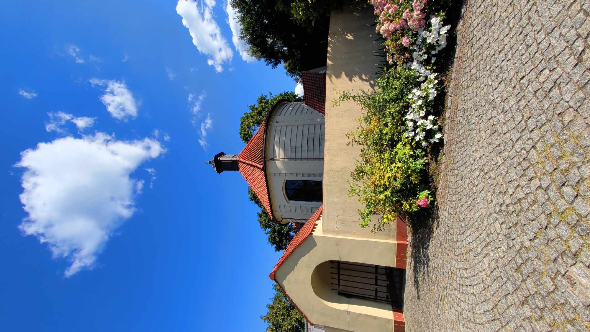 The image shows a circular building with a red-tiled roof and a black cupola, possibly a chapel or a mausoleum, situated behind a beige wall. An arched gateway with an iron gate is visible on the left side of the wall. In front of the wall, there is a garden with various flowering bushes, including white and pink flowers. The ground in the foreground is paved with cobblestones. The sky is bright blue with some white clouds. The building and garden are surrounded by trees. The scene appears to be a historical or religious site in the city of Pułtusk, Poland.