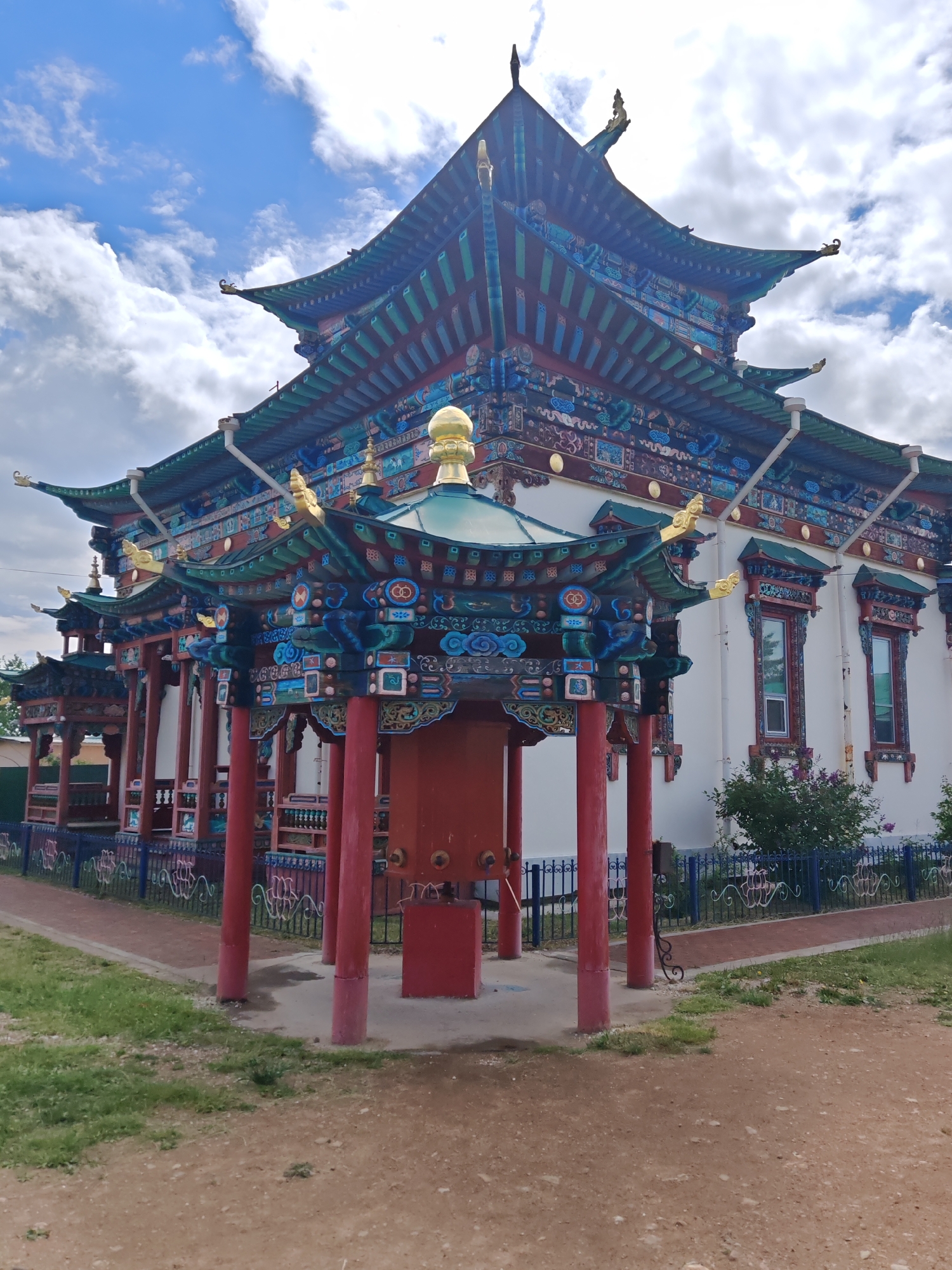 This media file showcases a vibrant, ornately decorated Buddhist temple or structure. The building features multiple tiered roofs with upturned eaves, painted in striking shades of teal, blue, and red, accented with gold embellishments. Intricate carvings and decorative patterns adorn the eaves and columns. Prominent red pillars support a covered walkway or porch in the foreground.
A large, cylindrical red object, possibly a prayer wheel, stands centrally within the foreground structure. The main building facade is painted white, with tall, narrow windows featuring decorative frames. A blue fence with a wrought iron-style pattern encloses the area, and a red path runs alongside it. Green grass and patches of dirt make up the ground around the building.
The sky above is a mix of blue with fluffy white clouds, suggesting it is daytime. The lighting indicates it might be late morning or early afternoon, with clear, bright conditions. The scene is serene and evokes a sense of cultural or religious significance, with no people visible. The location is not explicitly identified, but the architectural style suggests an East Asian influence, possibly in a rural or semi-urban setting. There is no visible text in the image.