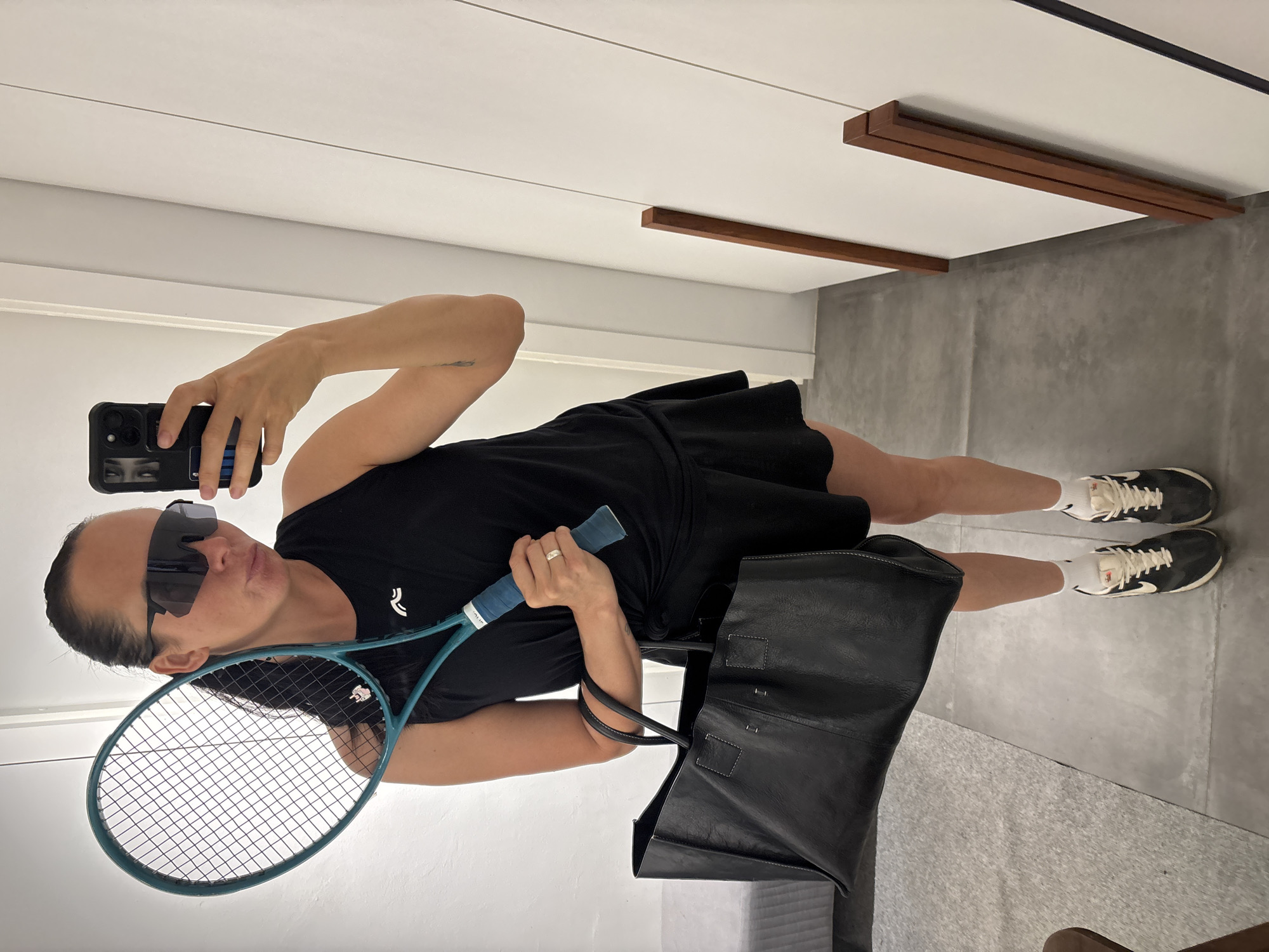 A woman is taking a selfie in what appears to be an interior setting. She is wearing sunglasses, a black athletic dress, white socks, and black and white sneakers. She holds a blue tennis racket in one hand and a black tote bag hangs from her shoulder. She is holding a phone in her other hand to take the picture. The background consists of a white wall with horizontal lines and some brown wooden accents. The floor is grey. The location in the city of Chaikovsky, Russia is unknown from the image.