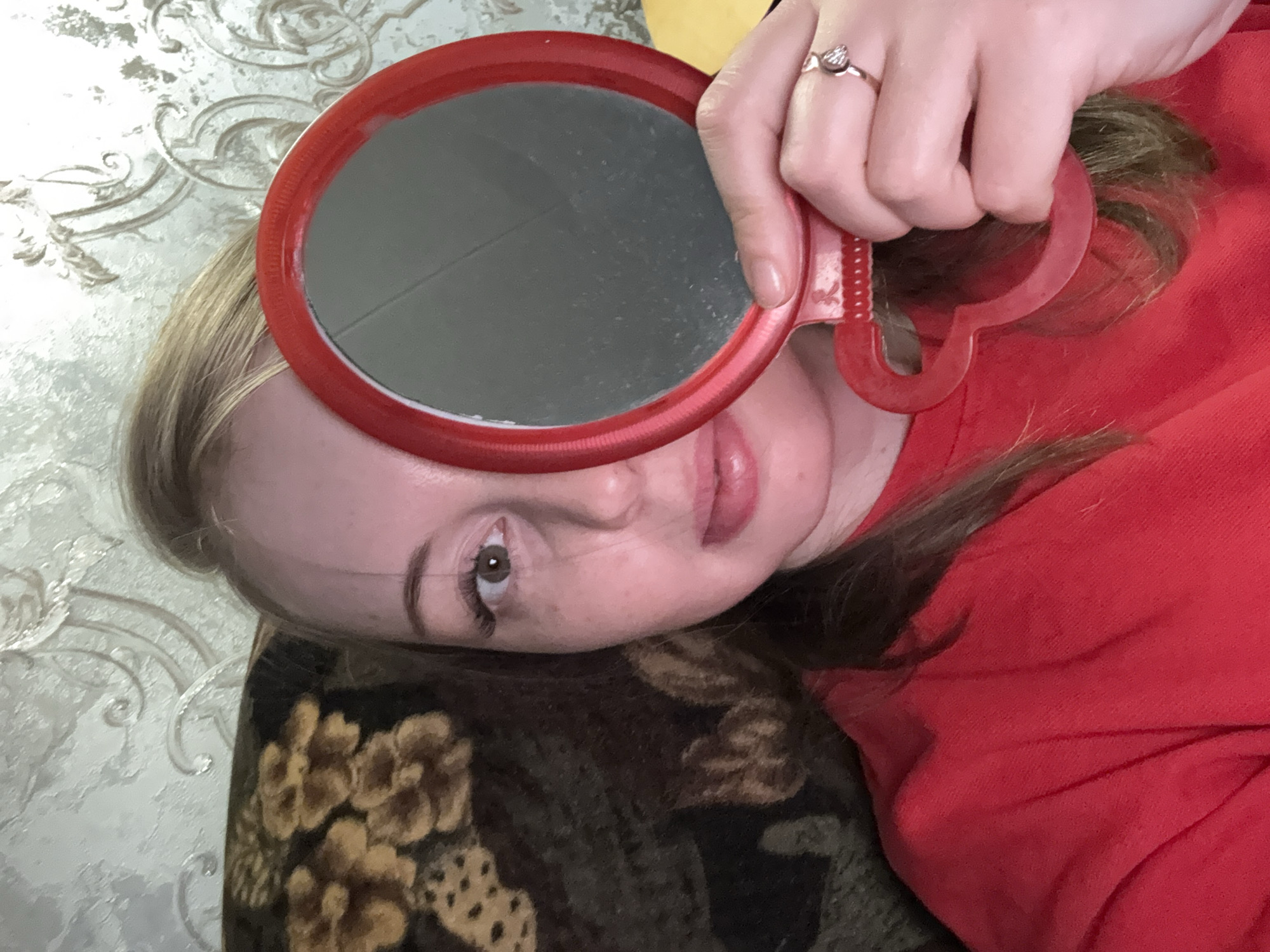 A medium close-up, tilted portrait shows a person, likely female, holding a red circular handheld mirror. The person's head is angled, with her right eye and the lower part of her face clearly visible. She has fair skin, light brown hair, and brown eyes, looking directly at the camera with a neutral expression. Her lips are adorned with pink lip gloss.
Her right hand, wearing a silver-colored ring with a prominent, possibly heart-shaped, clear stone on the ring finger, holds the red mirror near her face. The mirror's surface is largely grey with a distinct horizontal line or scratch across it, reflecting ambient light. The mirror partially covers her forehead and the left side of her face.
She is wearing a red garment, possibly a sweatshirt or sweater, visible on the right side of the frame. Below her head and to the left, a dark-colored piece of furniture, likely an upholstered couch or armchair, featuring a brown and gold ornate floral pattern, is visible. The background in the upper left corner shows a light-colored wall with textured, decorative wallpaper. The scene suggests an indoor, casual setting, possibly a home. There is no visible text in the image.