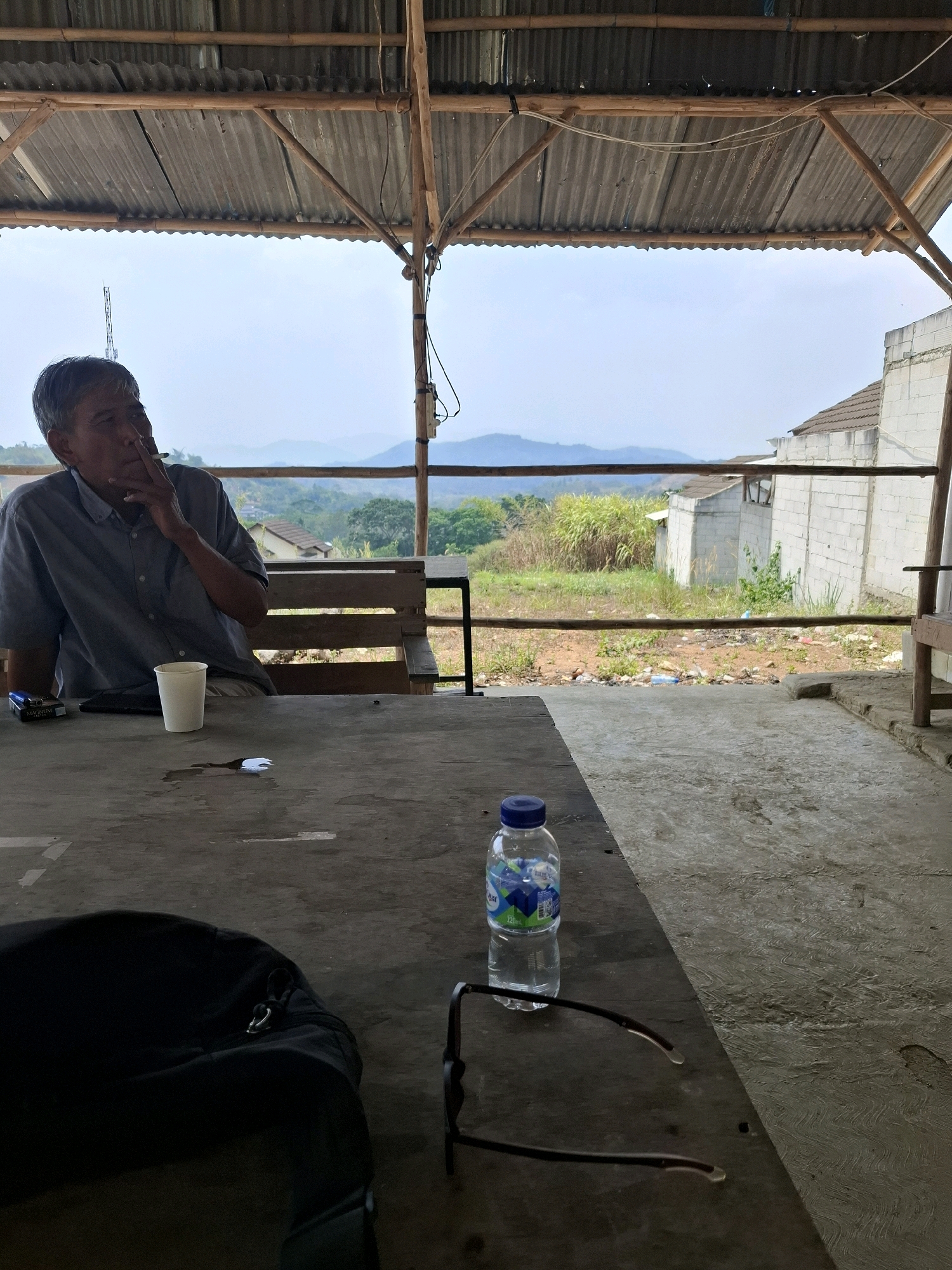 A man sits at a table under a corrugated metal roof supported by bamboo poles. He is wearing a blue collared shirt and is smoking a cigarette. In front of him is a white paper cup, a pack of cigarettes, and a cell phone. The table is made of weathered concrete. Beyond the table, a view of rolling hills and vegetation is visible, with some buildings in the distance. The ground is covered with debris. The man is the sole subject in the foreground. The setting appears to be an outdoor shaded area.