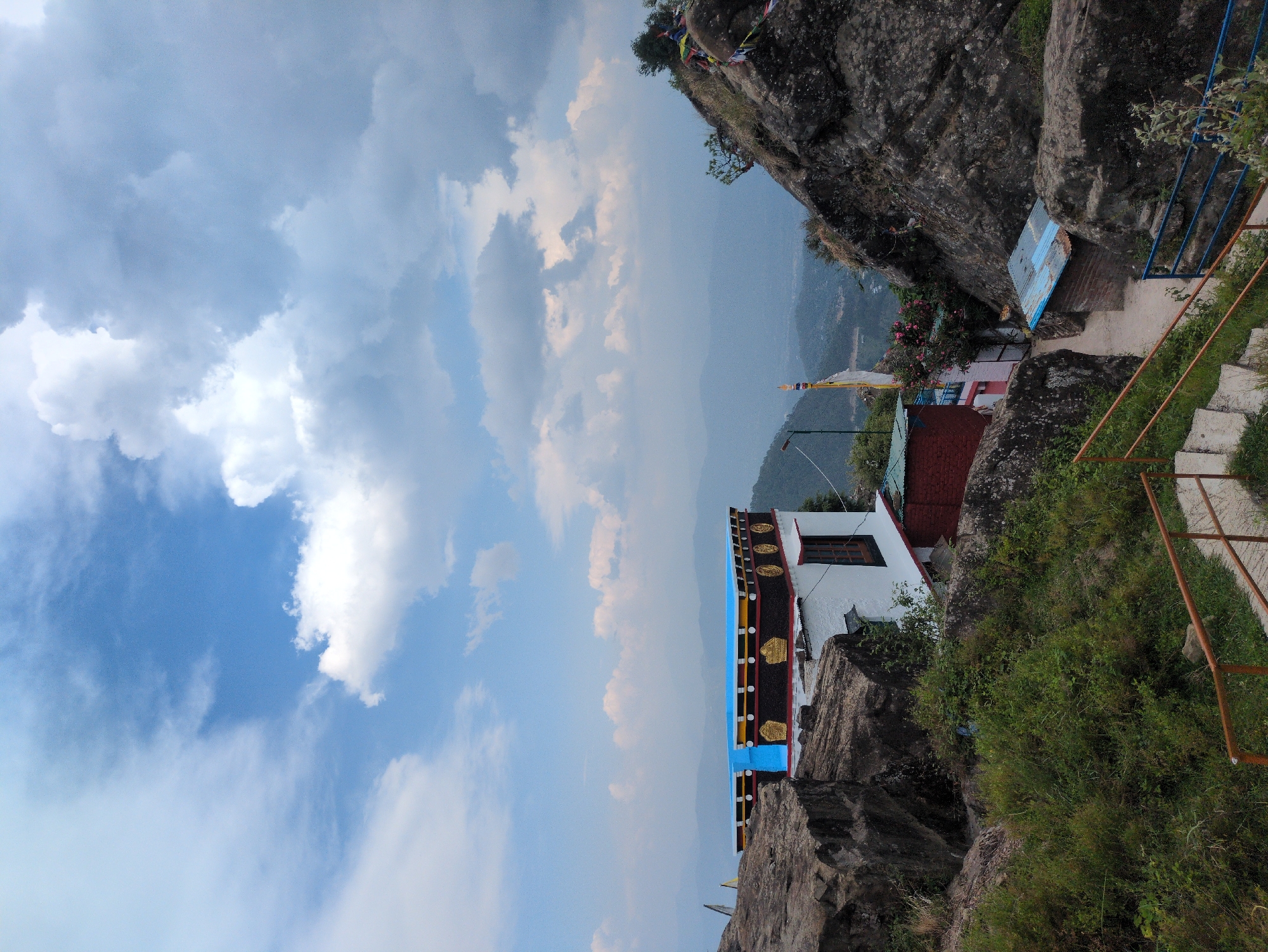 The image depicts a landscape scene with a building situated on a rocky incline, with mountains and sky in the background. The building is white with a colorful decorative roof, featuring blue, yellow, and black elements, and gold circular motifs. A wooden-framed window is visible on the side of the building. The surrounding environment is characterized by large rock formations and lush green vegetation. A pathway with railings leads down the incline. Several prayer flags are seen attached to rocks and poles. In the distance, a hazy view of mountains stretches across the horizon under a partly cloudy sky. The scene is located in the city of Doh, India.
