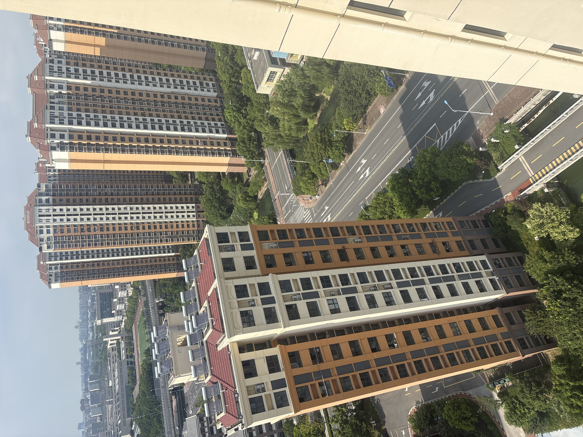 The image depicts a view of a residential area in Suzhou Industrial Park, China, featuring several tall apartment buildings. The foreground is dominated by a modern apartment complex with orange and cream colored facades, equipped with numerous windows and air conditioning units. On the rooftops of some buildings, solar water heaters are visible.
Beyond the immediate buildings, a multi-lane road runs through the scene, with cars and traffic markings. Lush green trees and foliage line the road and surround the buildings, with a canal or small body of water visible beneath a bridge. In the background, a sprawling cityscape of more apartment buildings and structures stretches towards the horizon under a hazy sky. The overall scene suggests a well-developed urban environment with a focus on high-density housing and infrastructure.