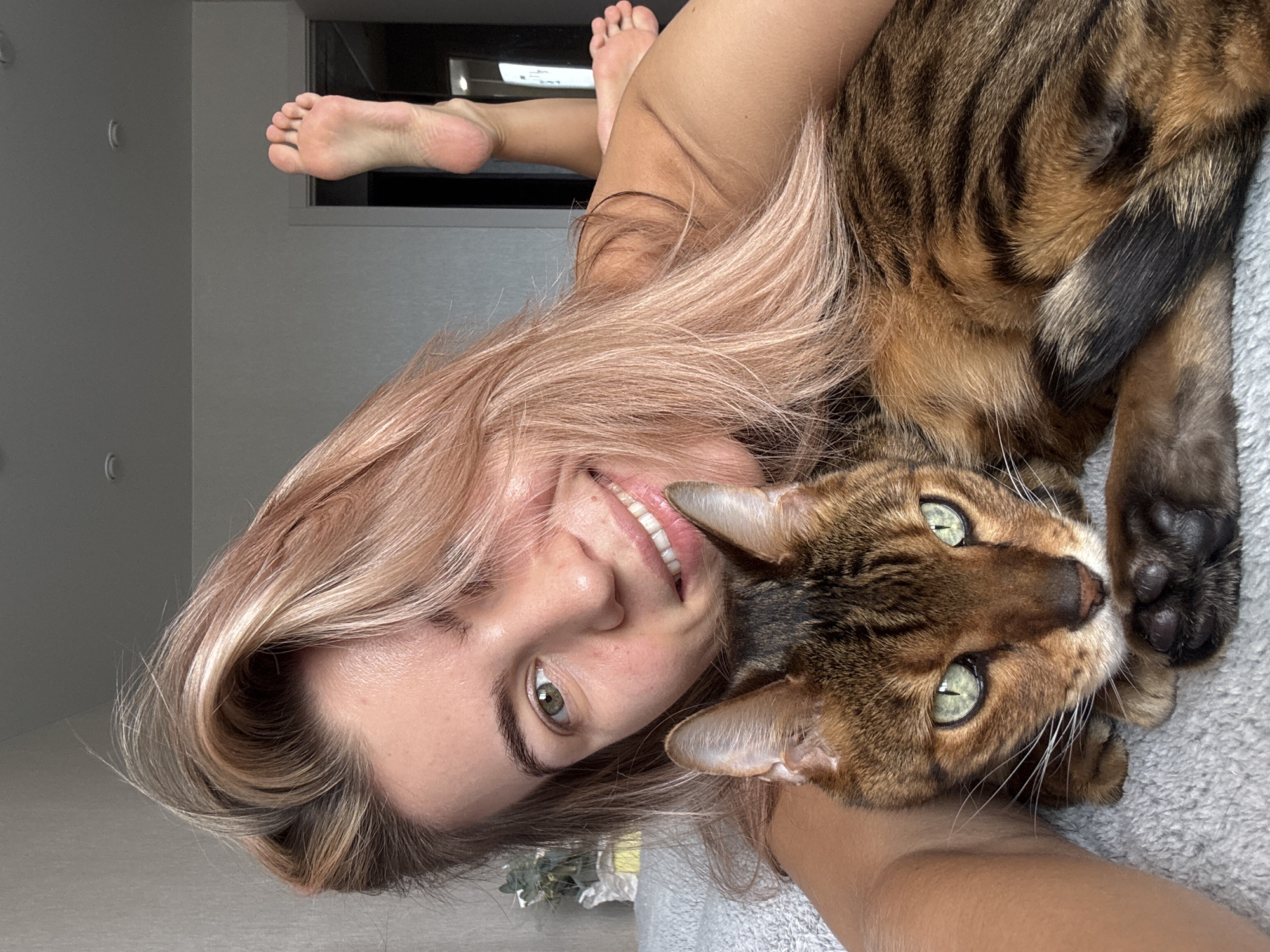 A woman and a cat are in the image. The woman has blond hair and is smiling. The cat is lying on the woman's chest and has green eyes. The woman is lying on a bed in a room with gray walls. There is a small window visible in the background. The location is Unknown, Unknown.