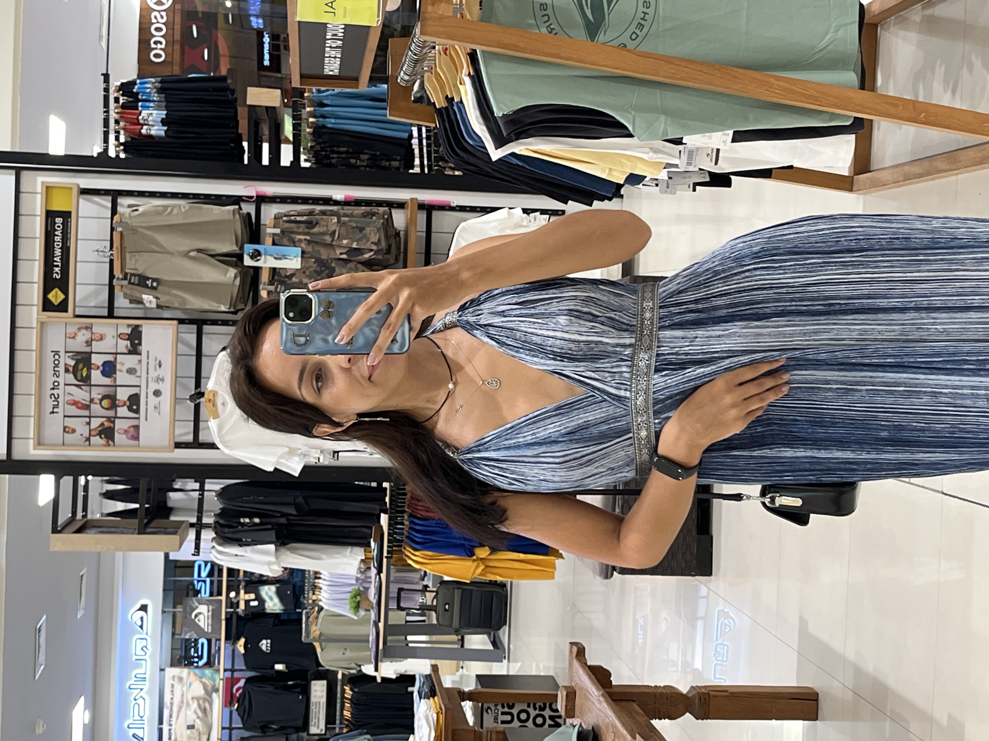 The image shows a woman taking a selfie in what appears to be a retail store, likely in a shopping mall in Unknown, Indonesia. The store sells clothing. The woman is wearing a blue, horizontally striped dress. She holds a smartphone in her left hand, taking the selfie. The background features racks of clothing, including t-shirts, shorts, and pants. Signage visible includes the word "BOARDWALKS" and the logo "ROCO." The environment is brightly lit, suggesting a typical commercial setting. A purse hangs on the left side of her arm.