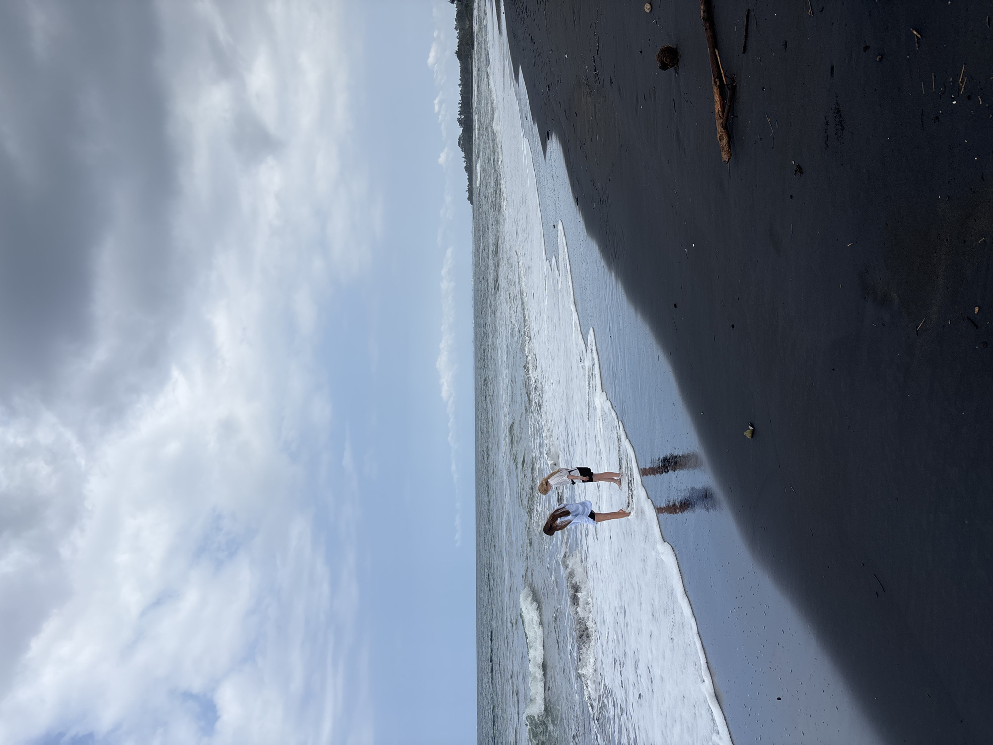 Two individuals, one with dark hair wearing a blue top and black shorts, and another with blonde hair wearing a white top and black shorts, stand at the edge of the water on a black sand beach. They are facing each other and appear to be interacting. The ocean is to their left, with waves breaking on the shore, creating white foam. The sky above is overcast with dramatic clouds. In the distance, a tree line is visible along the horizon. The scene is located on the beach in Blahbatuh, Indonesia.