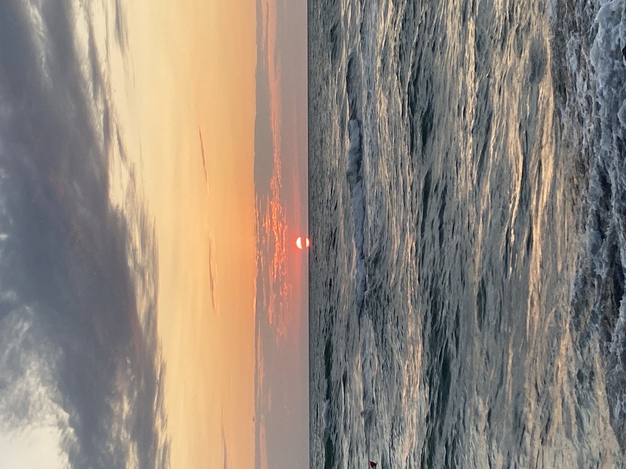 This is a photograph taken at sunset on the beach in Ureki, Georgia. The sun, a vibrant orange circle, is positioned just above the horizon, casting a warm glow across the sky. The sky is a gradient of colors, transitioning from soft oranges and pinks near the sun to darker grays and blues at the top, indicating gathering clouds. The sea is calm, with gentle waves rolling towards the shore. The water reflects the colors of the sunset, creating shimmering patterns. There are no visible people or specific structures in the image, emphasizing the natural beauty of the coastal landscape at dusk.
