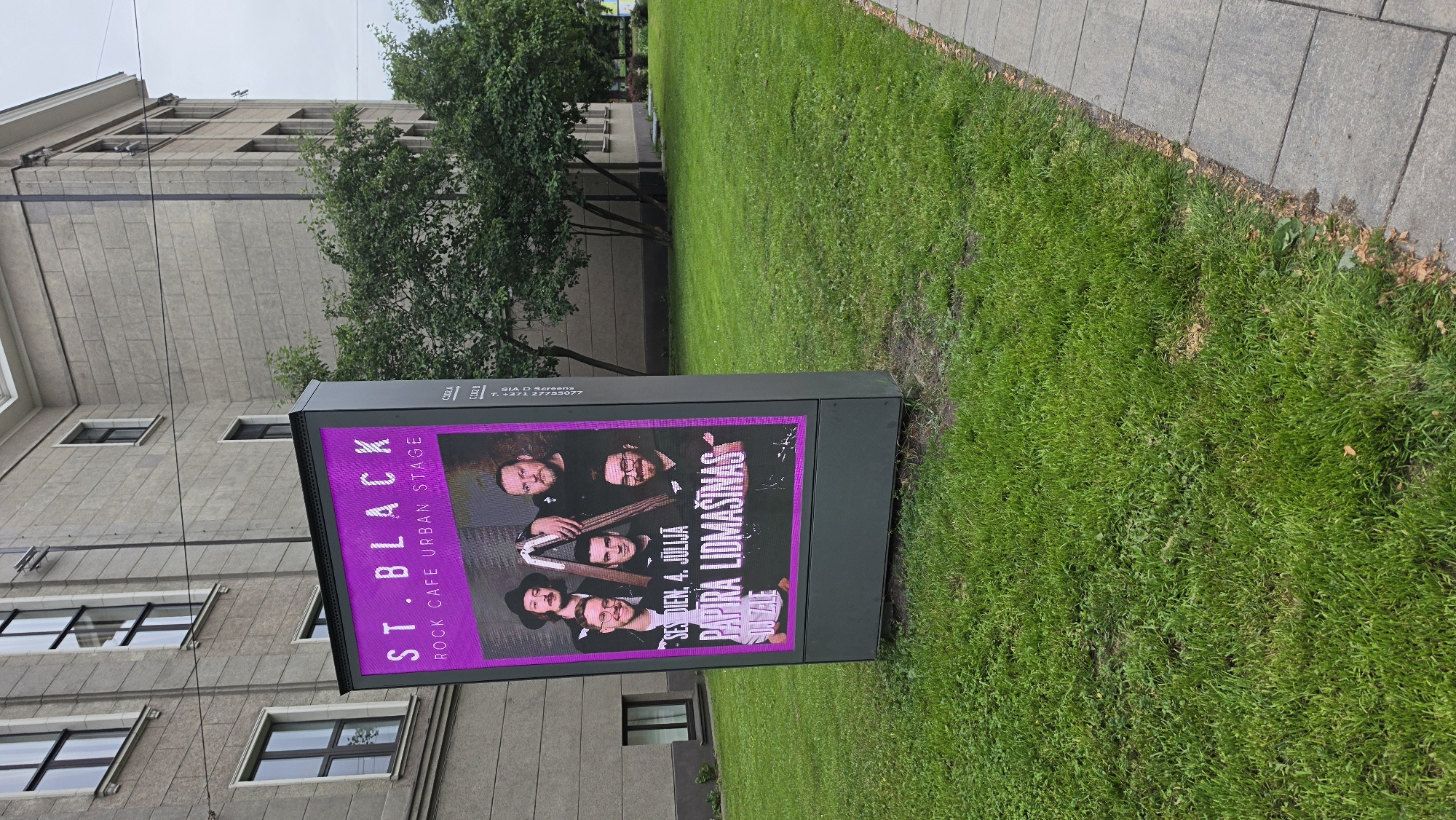 The image shows a poster displayed on a digital billboard outside a building with a grassy area and a sidewalk. The poster is advertising an event at "ST.BLACK ROCK CAFE URBAN STAGE". It features a photograph of a band of five men, likely musicians, posing together. The text on the poster indicates the event is on "SES DIEN, 4. JŪLIJĀ" which translates to "Wednesday, July 4th". Below the band's name, it also mentions "PAPIRA LIDMAŠĪNAS" and "DJ ZALE", suggesting a performance by the band Papīra Lidmašīnas with a DJ named Zale. The billboard is dark grey and positioned on a patch of green grass next to a grey tiled sidewalk. In the background, part of a building with multiple windows is visible. The sky appears overcast, suggesting it could be a cloudy day. The overall impression is an advertisement for a live music event at a rock cafe.