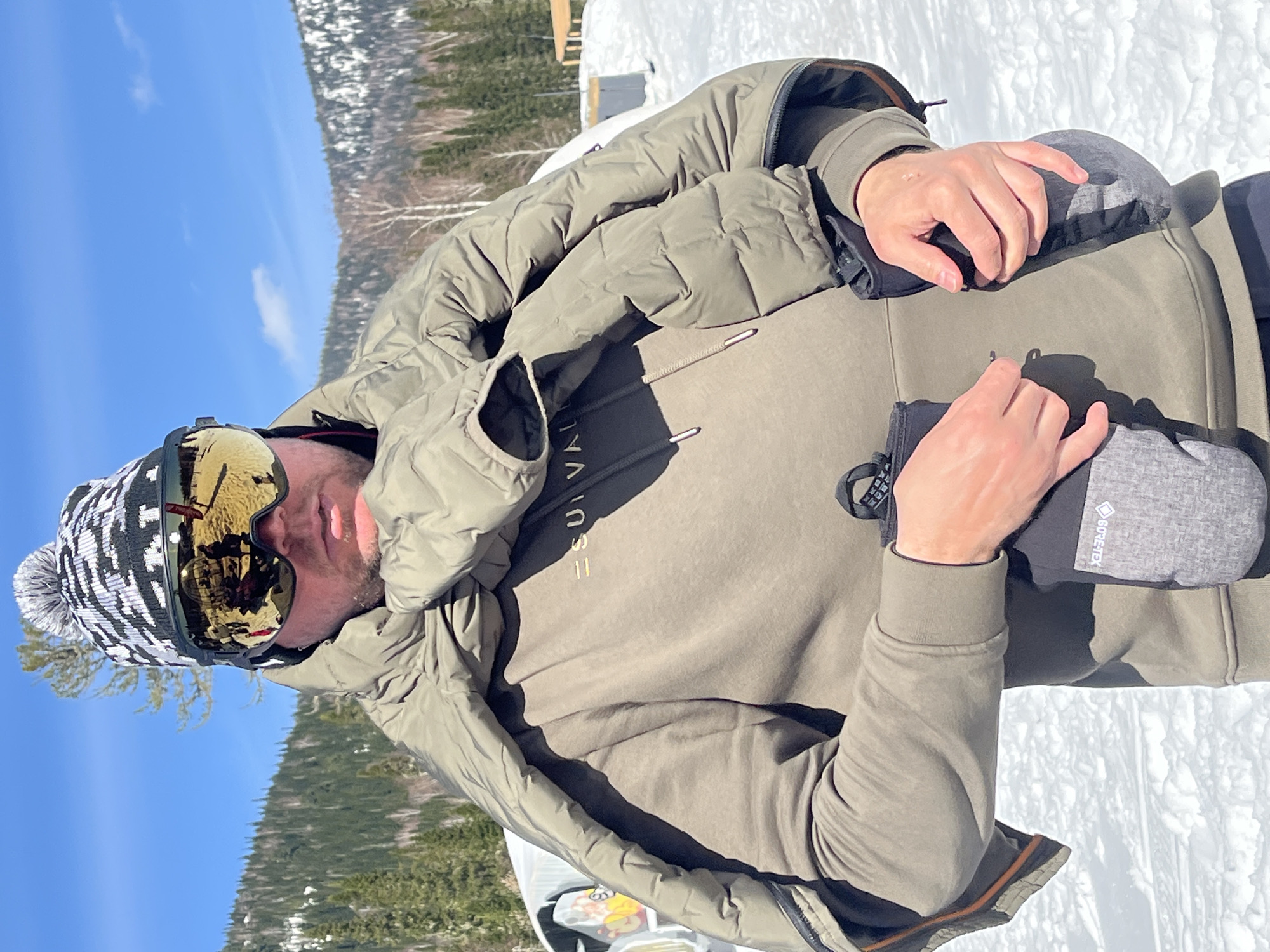 A man is standing in a snowy, mountainous outdoor environment in an unknown location. He is wearing a black and white knit hat with a pom-pom, gold-tinted ski goggles, an olive-colored sweatshirt with an indiscernible inscription across the chest, and a puffy olive-colored jacket slung over his shoulders. He is holding a gray Gore-Tex glove in each hand. The background features a snow-covered landscape, including evergreen trees and a partly cloudy blue sky.