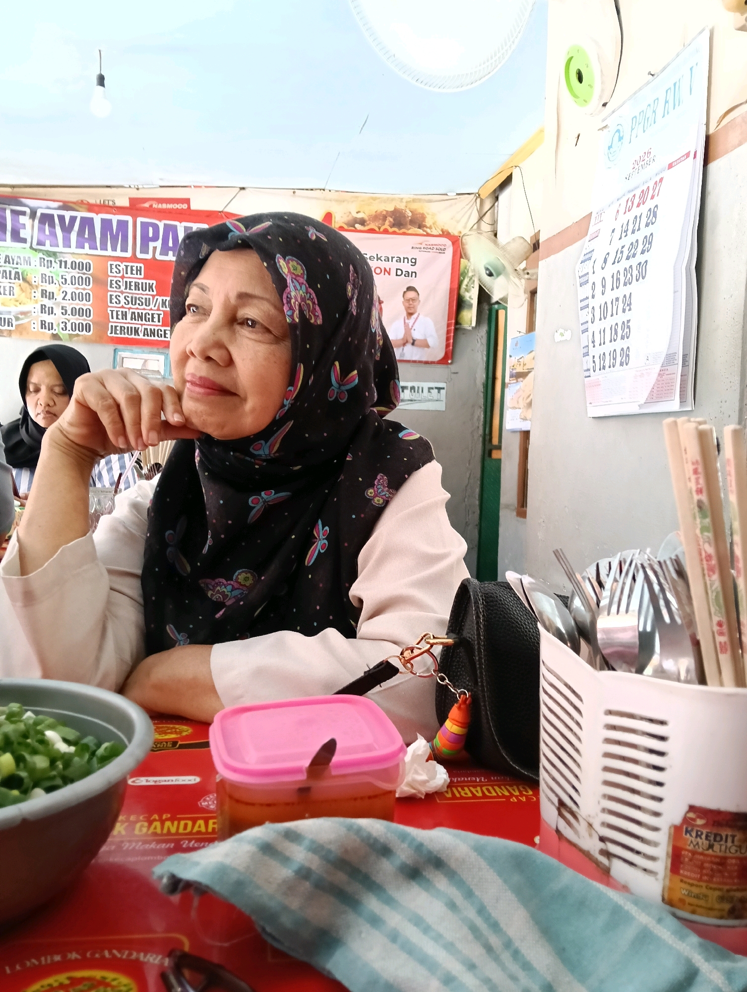 An outdoor, casual restaurant setting is depicted. The focal point is a woman wearing a black hijab and a light-colored blouse, with her hand resting on her chin, looking to the left of the frame. Behind her, another woman in a black hijab is visible. The background includes a banner with Indonesian text and an image of a man, a calendar with dates, and a stall selling food. On the table in the foreground are a bowl of green onions, a pink container of sauce, a blue and white striped cloth, a black purse with a colorful charm, and a cutlery holder filled with spoons and forks, as well as chopsticks. A small can with red and yellow labeling is also on the table. The overall lighting suggests daylight.
