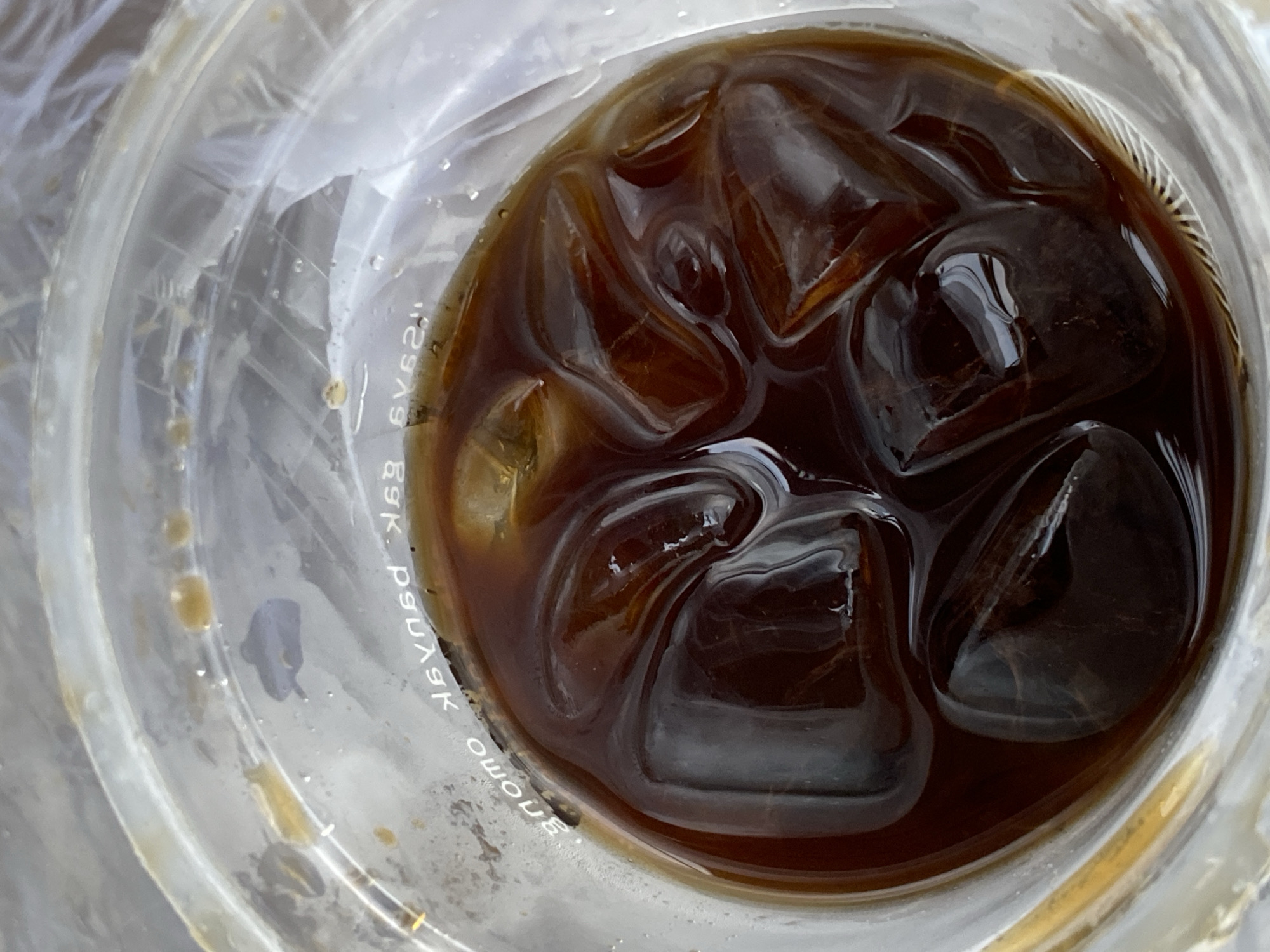 This is a top-down, close-up shot of iced coffee in a clear plastic cup. The coffee is dark brown and fills the cup, with several ice cubes submerged. There are some water droplets and condensation on the inside of the cup. Text is visible around the rim of the cup, but it is partially obscured by the coffee and ice, and appears to be in reverse. The background is blurry and out of focus, suggesting the cup is on a table or other surface. There is no indication of the city of Mengwi, Indonesia, or any other specific location.