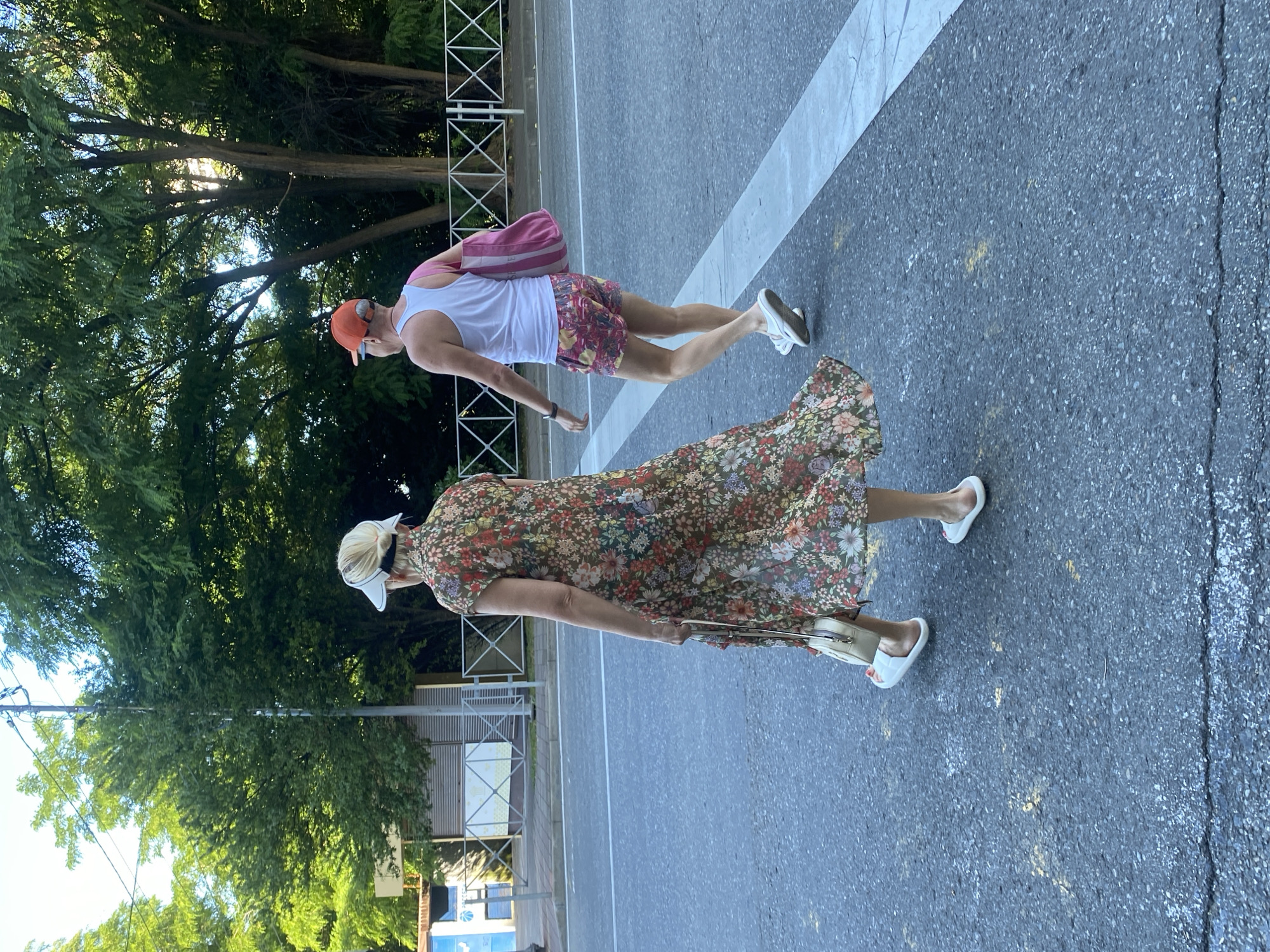 Two individuals are crossing a street in Sochi, Russia, on a paved road with white lane markings. The person on the left, viewed from behind, is wearing a floral print maxi dress, a white visor, and white slides, carrying a cream-colored handbag. The person on the right, also viewed from behind, is wearing a white tank top, colorful patterned shorts, an orange baseball cap, and white sneakers, and is carrying a pink and white striped tote bag over their right shoulder. They are walking from the right side of the frame towards the left. Behind them, trees with lush green foliage line the street, and a white metal fence is visible. In the background, there are some buildings and a utility pole. The lighting suggests it is daytime, possibly late afternoon, with sunlight filtering through the trees.