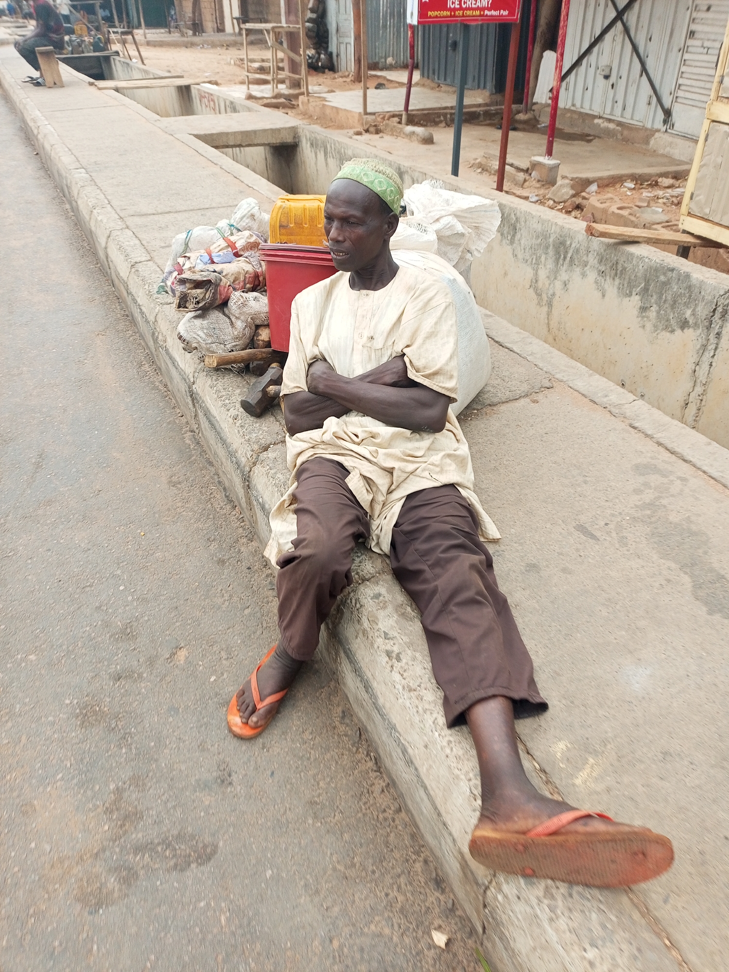 An elderly, dark-skinned man is seated on a concrete curb alongside a paved road in Tempure, Nigeria. He wears a light-colored, long-sleeved tunic (agbada-style), dark brown trousers, and bright orange flip-flops. A light green, patterned skullcap adorns his head. His arms are crossed, and he leans slightly against a large white bag, with a neutral, possibly pensive or weary expression on his face.
Next to him, on the curb, are several bundled white bags, a red plastic crate, a yellow jerry can, and two hammers lying on the ground. These items suggest he might be a vendor, a laborer, or someone traveling with personal belongings.
The setting appears to be a semi-urban or rural roadside. A concrete gutter system runs parallel to the curb, separating the road from an unpaved area with various simple structures and buildings, some with corrugated metal walls. In the background, a red sign with white text is visible, reading "ICE CREAM?" and "POPCORN + ICE CREAM = Perfect Pair," indicating a nearby refreshment stand or shop. The lighting suggests it is daytime, possibly overcast or late afternoon, with no harsh shadows. The man appears to be resting or waiting by the roadside.