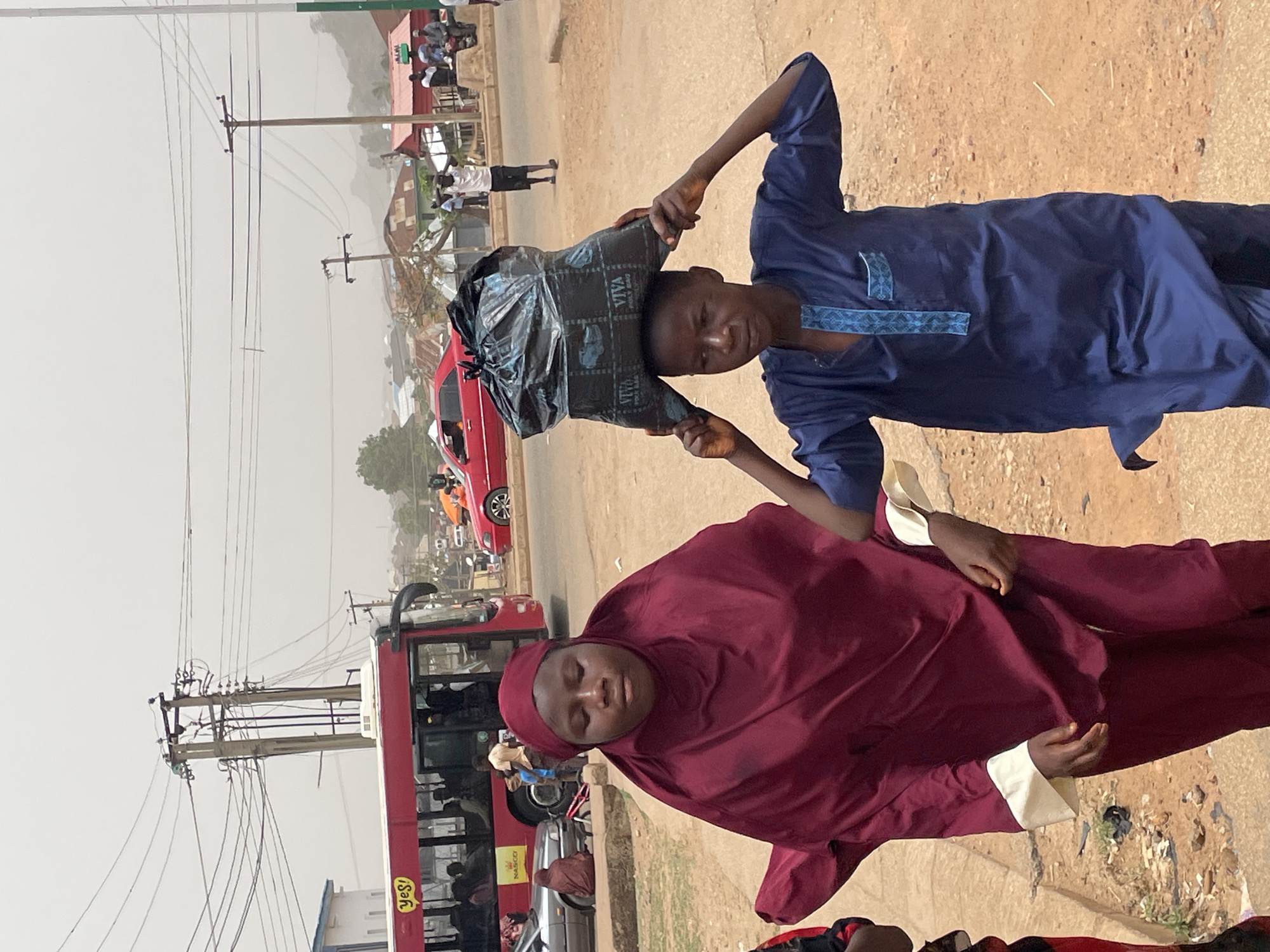The scene depicts a bustling street in Jos, Nigeria, under a hazy sky with overhead power lines. A young man balances a black plastic bag, marked "VIVA," atop his head, using his hands for support. He wears a dark blue traditional outfit with light blue patterned accents. Beside him, a woman in a maroon hijab with white cuffs stands. In the background, a red bus with visible passengers and logos is present, as well as other cars, buildings, and pedestrians. The ground appears to be a combination of paved road and dusty terrain.