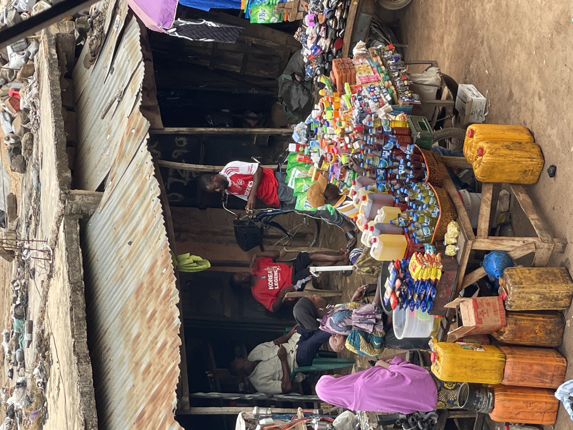 The image, taken in Bukuru, Nigeria, shows a street scene dominated by a densely packed informal market stall. The stall is built with corrugated iron and rough wooden supports. It is piled high with various goods, including colorful bottles, containers of liquids (some in yellow jerrycans), packaged items, and other miscellaneous merchandise.
Several people are present. One man, wearing an Arsenal soccer jersey, appears to be fixing a bicycle next to the stall. Another man wearing a red t-shirt, is resting on a chair. Further back, more people are sitting and lying down within the structure of the market.
The surrounding environment includes a rough, unpaved ground surface and adjacent dilapidated structures and assorted materials. The scene suggests a crowded and economically active area.