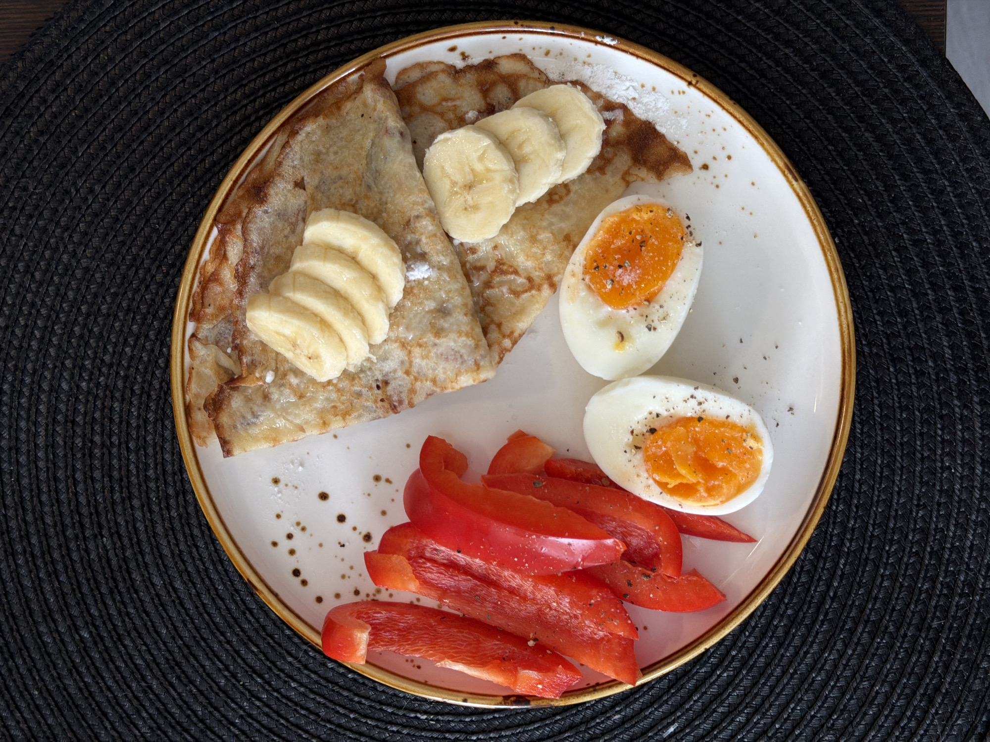 A high-angle, medium shot depicts a plate of food on a dark, textured surface, possibly a placemat. The plate contains two crepes, sliced bananas, hard-boiled eggs, and sliced red peppers. The crepes appear to be dusted with powdered sugar. The eggs are halved and sprinkled with pepper. The plate has a white center with a brown rim. The food composition suggests a breakfast or brunch meal. The location is Засечное, Russia.