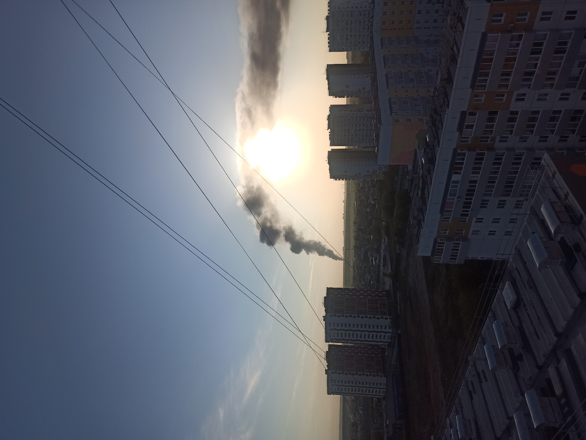 The image, likely taken from a high vantage point, shows a scene in the urban area of Кондратово, Russia. The photograph captures a line of densely packed apartment buildings oriented horizontally across the right side of the frame. In the background, rising from the ground, is a column of dark gray smoke. The smoke appears to originate from an area behind the apartment buildings. The sun is obscured by a thinner part of the smoke cloud. Three parallel power lines stretch across the left half of the image and bisect the sky and the smoke. The sky shifts from dark blue in the upper left corner to a light yellow hue near the sun. In the bottom right corner, many small rectangular shapes suggest a storage or industrial area.