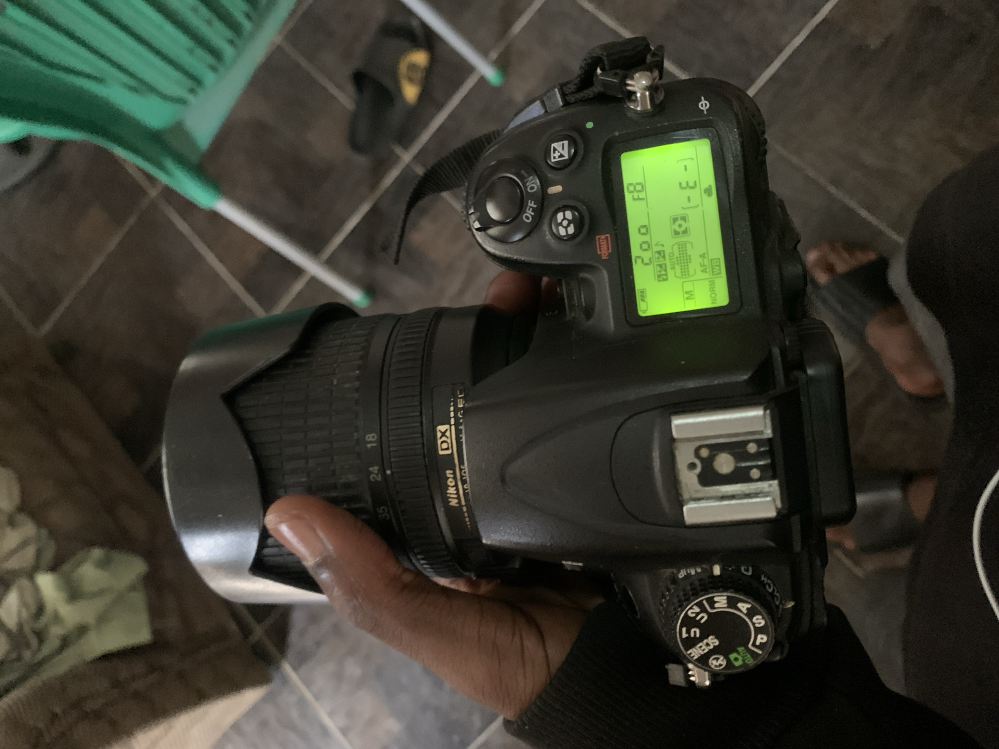 The image shows a close-up, top-down view of a person holding a black Nikon DSLR camera. The camera has a lens attached with the brand name "Nikon DX" visible on it, along with focal length markings including 18, 24, and 35. The camera's LCD screen is illuminated and displays settings including "200", "F8", "AUTO", "AF-A", "NORM", and "WB". A mode dial on the top of the camera shows various settings like "AUTO", "P", "S", "A", "M", "U1", "U2", and "SCENE".
The background of the image suggests an indoor setting. A portion of a green plastic chair is visible in the upper left corner. The floor appears to be tiled with a dark grout. A pair of black flip-flops is partially visible in the upper center of the image. The lighting in the image appears to be artificial. The person's hand holding the camera is in focus, indicating the photographer's perspective. The overall impression is of someone preparing to take a photograph or examining their camera settings in a casual environment. No specific time of day or weather conditions can be determined. The location context provided indicates Jos, Nigeria.