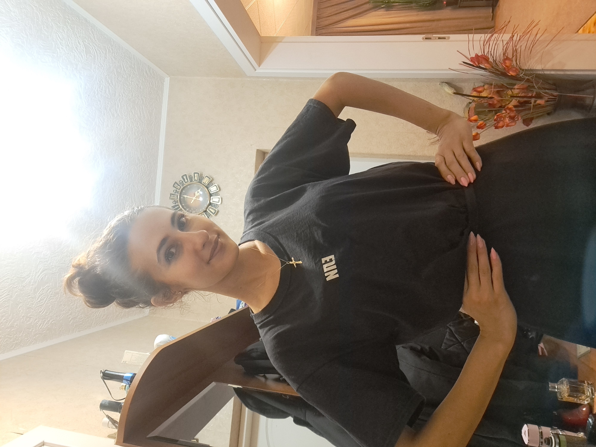 The media file shows a young woman posing for a picture indoors, possibly in a bedroom or living area. She is wearing a black t-shirt with a small gold cross necklace and the word "FUN" printed on the shirt in white lettering. Her hair is pulled up in a messy bun, and she is looking at the camera with a slight smile. Her left hand is placed on her hip, and her right arm is bent at the elbow. In the background, there is a decorative clock on the wall, and a vase with artificial flowers. A wooden shelf is also visible, with some items on it that are not clearly identifiable. The lighting suggests it is daytime, with a bright light source coming from the left, possibly a window or a strong lamp. The overall mood of the image is casual and relaxed.
