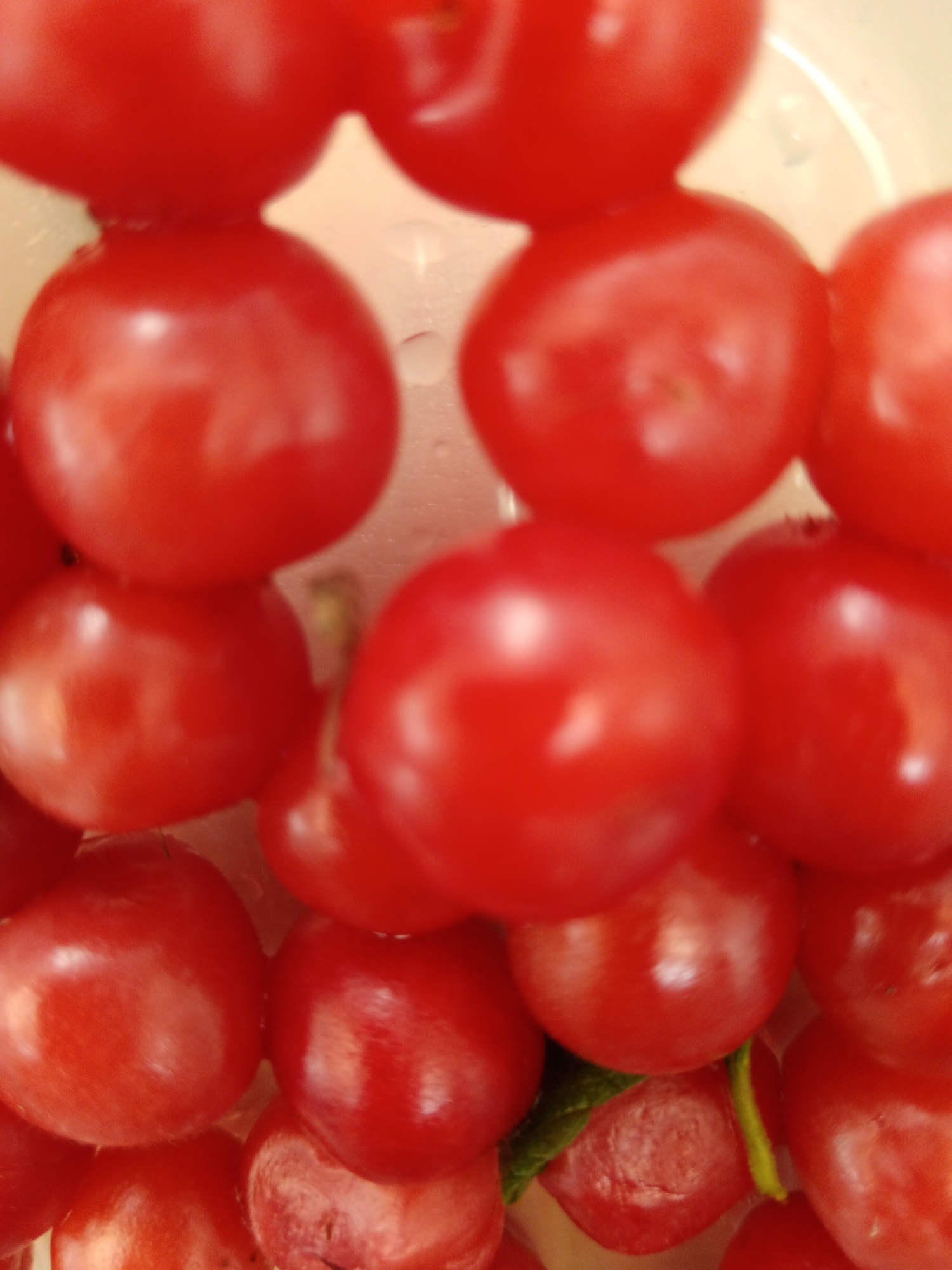 A close-up, overhead shot features a cluster of ripe, red cherry tomatoes. The tomatoes are arranged in a way that suggests they are piled on top of each other, with some partially obscuring others. The lighting highlights the glossy texture of the tomatoes, with soft reflections visible on their surfaces. A hint of green, possibly a stem or leaf, is visible near the bottom center of the image, attached to one of the tomatoes. The background is a muted, pale color, possibly white or cream, which makes the vibrant red of the tomatoes stand out. There are no people or other objects visible in the frame. The scene is likely a domestic setting, perhaps a kitchen counter or a bowl used for displaying produce. It is not possible to determine the location in Sorochinsk, Russia from the image.