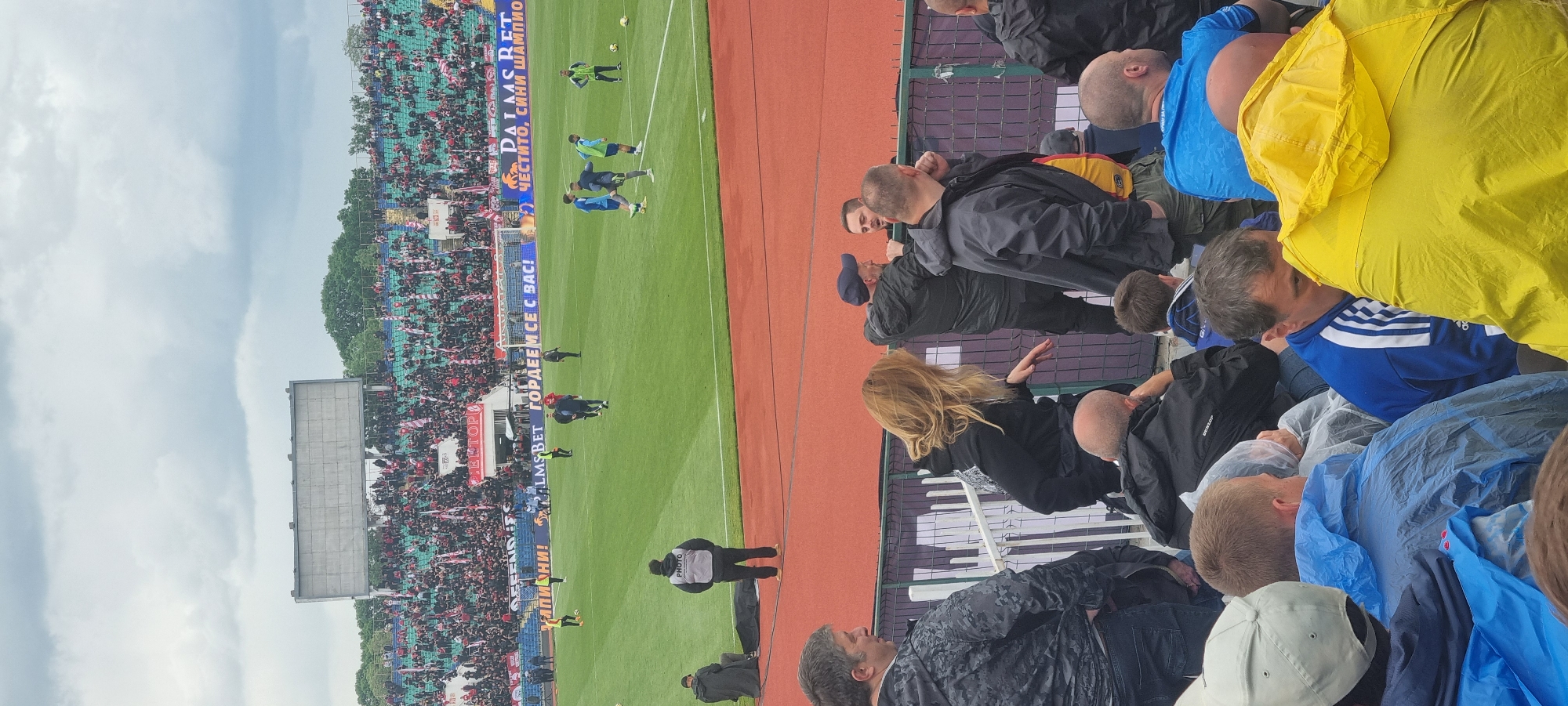 The image captures a soccer game being played in a stadium, likely outdoors on a partially overcast day. A segment of the audience is visible on the right side of the frame, composed of individuals wearing an array of clothing, including a bright yellow raincoat and a blue jacket with white stripes. Some people are seen wearing plastic ponchos, suggesting rainy weather. On the left side of the image, spectators fill the stands, some waving banners or flags. The soccer field is green, with players in blue and green uniforms. A banner with printed text in what appears to be a Cyrillic alphabet is strung along the sideline. In the lower-left and central parts of the image, stadium security personnel are positioned along the edge of the field.