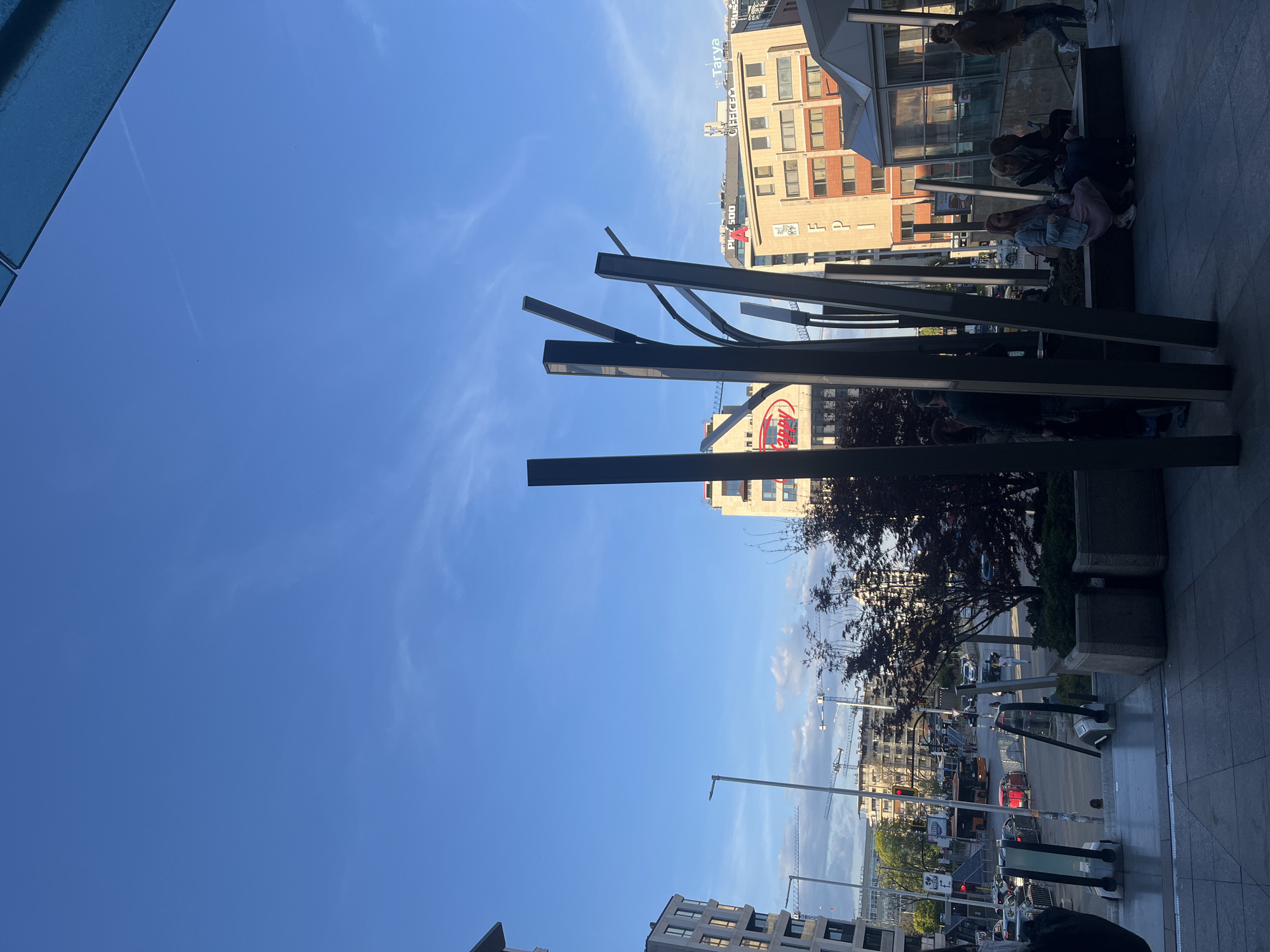 An outdoor urban scene in Sofia, Bulgaria, is captured under a bright, clear blue sky with sparse wispy clouds. The image appears to be taken from a slightly elevated or recessed perspective, looking out onto a street and a public plaza area.
Prominent in the foreground and midground are dark, abstract metal structures, resembling stylized, angular beams or architectural supports, which partially frame the view. To the upper left, a reflective blue surface, possibly part of a glass canopy or building facade, is visible.
The midground features several multi-story buildings. On a building to the right, the word "Tarva" and "PPC" are visible. Below it, another building displays a red circular logo with the text "SUD." Further to the right and slightly lower, a large red "Koka" logo (reminiscent of the Coca-Cola brand) is displayed on another building.
A group of people is gathered on a raised platform or seating area to the right, some seated and others standing. One person in the group is wearing a headscarf. They appear to be engaged in casual activity, such as relaxing or waiting.
In the background, a street is visible with moving vehicles, including cars and a larger truck, and functioning traffic lights. Several tall construction cranes are silhouetted against the sky in the distance, indicating urban development. Trees and planters with greenery are interspersed throughout the scene. The clear daylight casts distinct shadows, suggesting a sunny day, likely late afternoon or early morning.