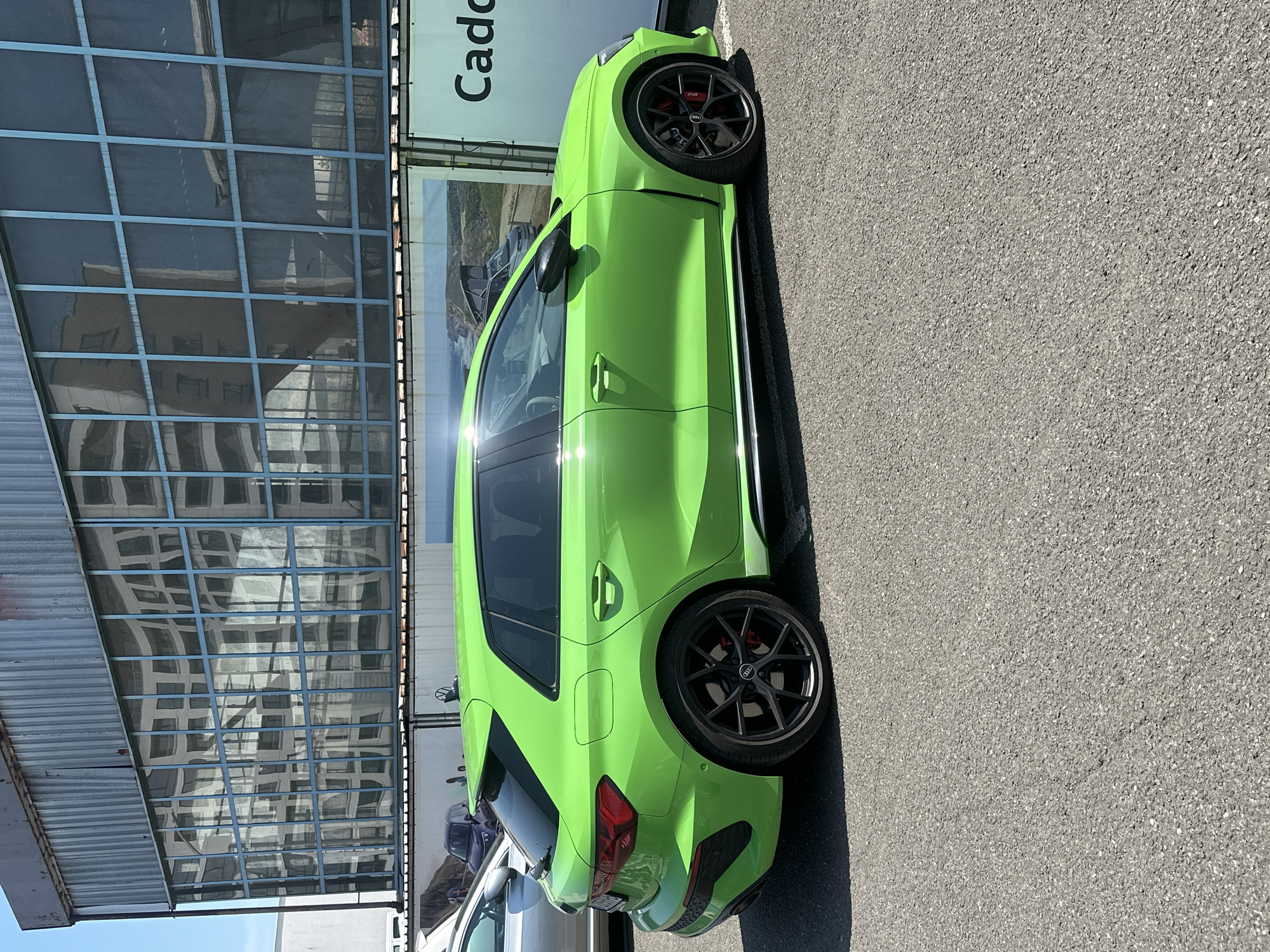 A vibrant, neon green Audi RS3 Sportback is the central focus of the image, parked outdoors on an asphalt surface under bright daylight. The car is positioned horizontally across the frame, showcasing its sleek side profile, rear three-quarter view, and a portion of its front. It is equipped with distinctive black multi-spoke alloy wheels, behind which red brake calipers bearing an "RS" logo are visible.
In the background, a large building with a contemporary, possibly industrial, design is visible. The upper section features extensive reflective glass windows arranged in a grid pattern, mirroring the surrounding environment, including a glimpse of a blue sky and other structures. Below this, a section of corrugated metal roofing is apparent. Adjacent to the building stands a large white sign with the prominently displayed word "Cado" in dark lettering. A large image, possibly an advertisement depicting a landscape scene with another vehicle, is also present on the sign.
The bright natural sunlight creates strong shadows on the asphalt beneath the car and generates noticeable glare and reflections on the car's glossy green paintwork and windows. The clear reflections in the building's windows suggest a sunny day with a blue sky. A light-colored vehicle is partially visible parked behind the green Audi. The overall setting appears to be a commercial or industrial area, such as a car dealership or a large parking facility.