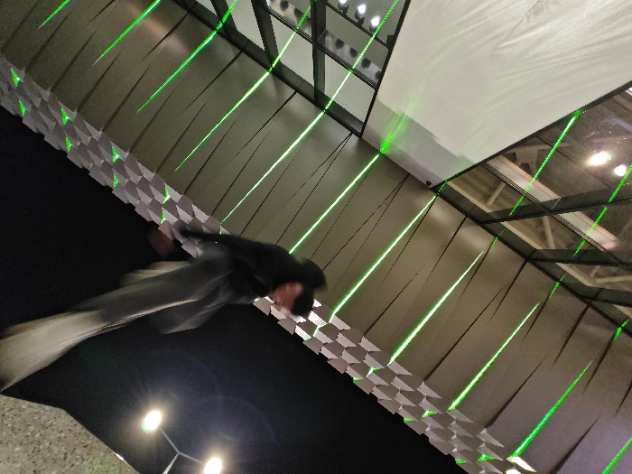 An indoor scene within Jump Park Shumen, Bulgaria, depicts a dynamic environment primarily lit by numerous diagonal green laser beams. The upper section features a ceiling structure composed of dark support beams and large, light-colored panels, with several bright light fixtures visible. Below the ceiling, a distinctive wall segment presents a repeating geometric pattern of white and grey angular blocks. In the lower-middle foreground, a single individual is captured in a state of significant motion blur, suggesting a rapid movement such as running or jumping. The person wears dark clothing on their upper body and lighter-colored pants, with their head partially obscured and angled towards the right. The lower ground level is predominantly dark, with additional light sources faintly visible in the bottom-left corner.