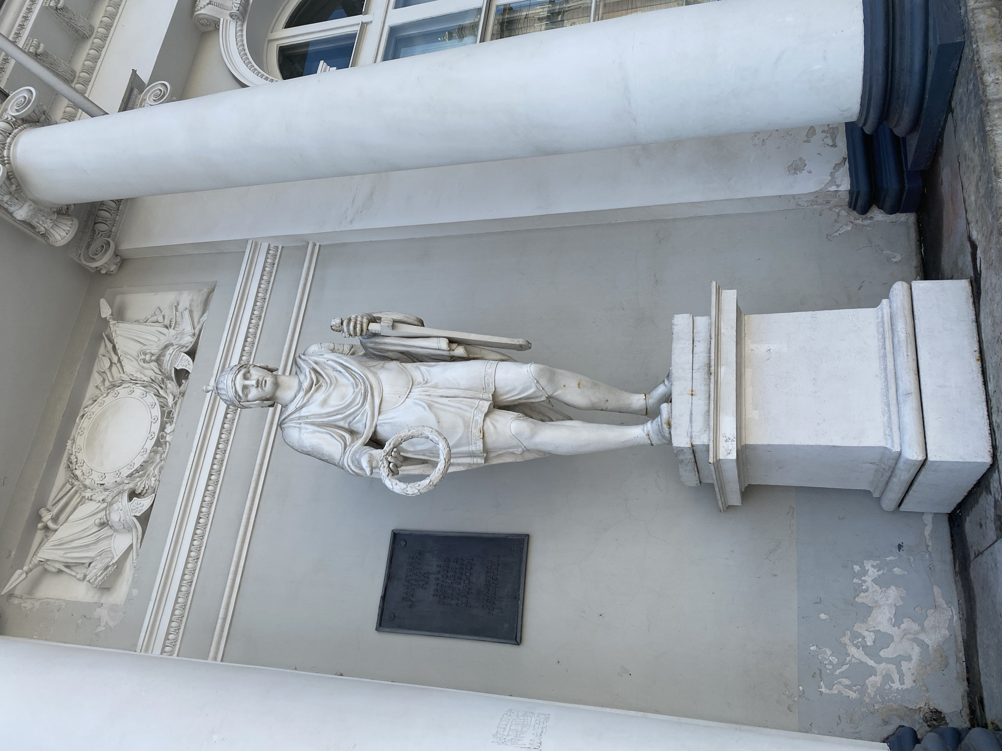 This image shows a white marble statue of a man in ancient attire, standing on a pedestal against a light gray wall. The statue is positioned in front of a classical building with large white columns and arched windows. A dark plaque is mounted on the wall to the left of the statue. The building appears to be located in Saint Petersburg, Russia. The statue is holding a laurel wreath in its left hand and a staff or scepter in its right. The wall behind the statue shows signs of weathering and peeling paint. The overall scene suggests a public square or the facade of a historical building.