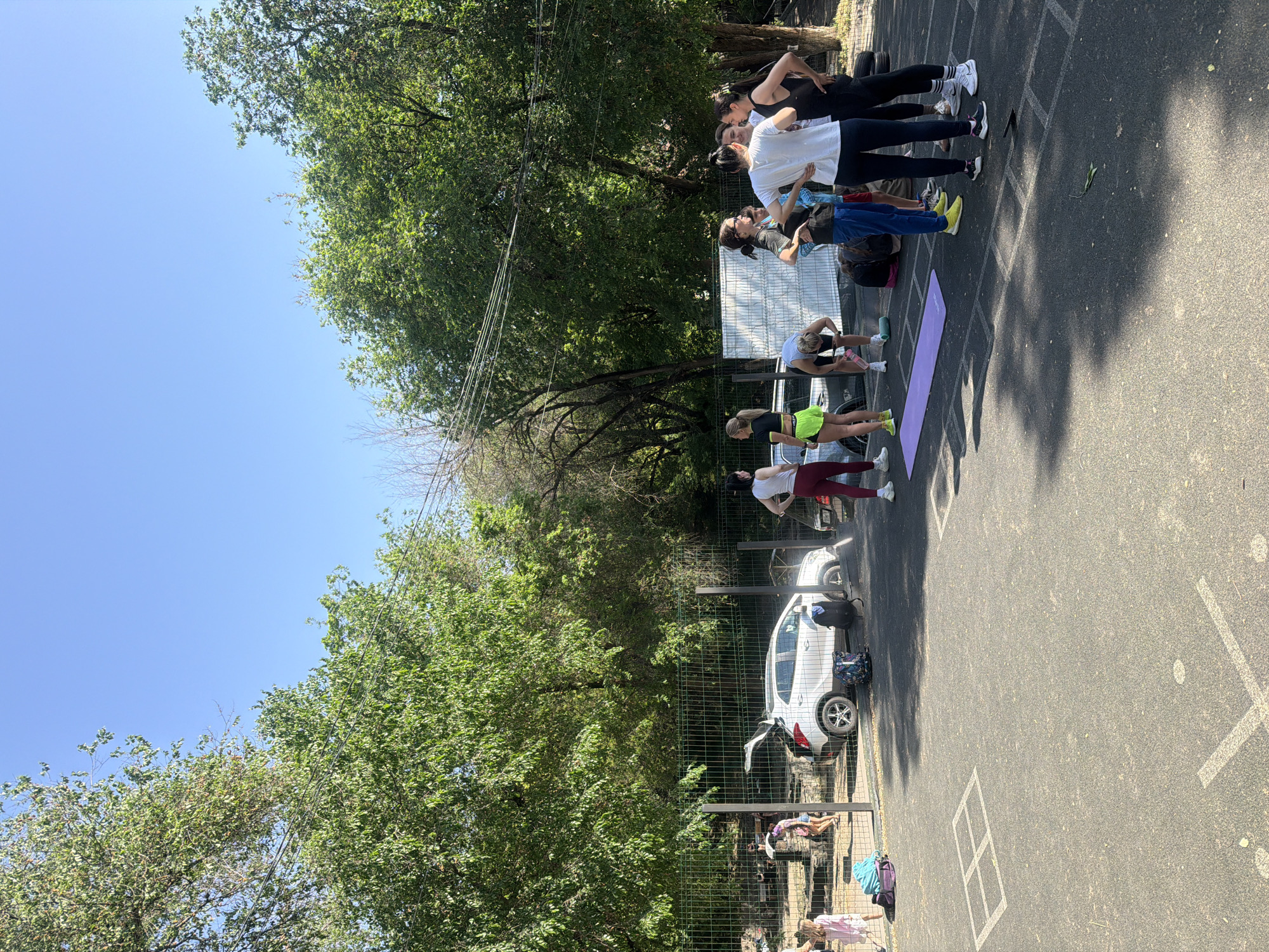 A group of women are exercising outside on an asphalt surface marked with white lines forming game patterns. They are stretching and bending over in a line formation. A white car with an open trunk is parked nearby. In the background, there is a green metal fence, trees, and a clear blue sky. A lower level area near the fence seems to be a playground with children and personal items scattered. This picture was taken in Rostov-on-Don, Russia.