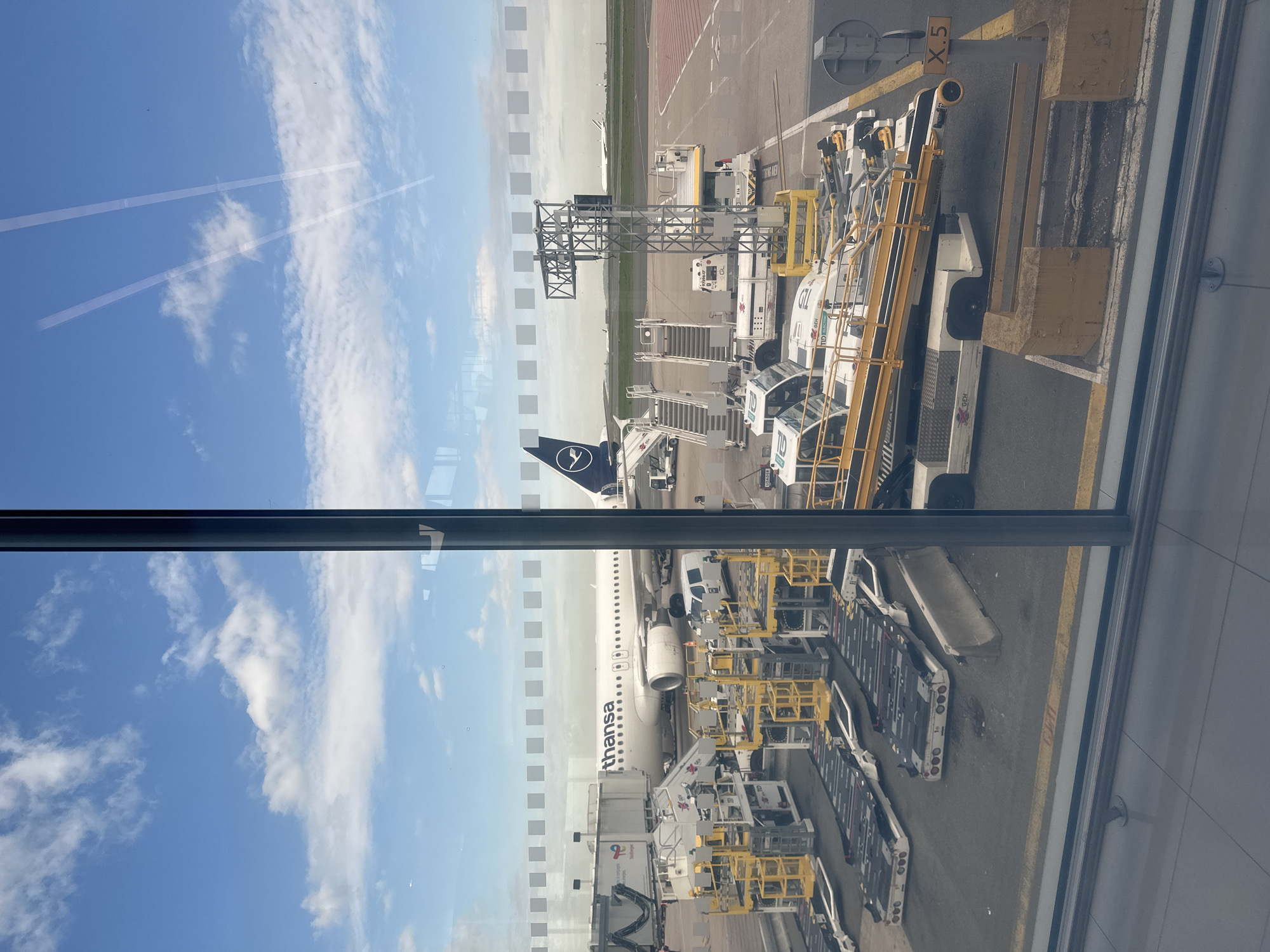 A daytime photograph taken from an airport terminal window shows a Lufthansa airplane parked at a gate on the tarmac, under a bright blue sky with scattered white clouds and visible contrails. The image is oriented vertically, showing the ground to the right and the sky to the left.
The white fuselage of a Lufthansa aircraft, with "Lufthansa" text prominently displayed along its side, is connected to a jet bridge. The blue tail fin, bearing the distinctive Lufthansa crane logo, is visible toward the upper left of the aircraft. Around the plane, various pieces of ground support equipment are positioned, indicating active airport operations. These include several white and yellow baggage carts or dollies, some marked with "GEH," ground service tugs, and mobile passenger stairs.
On the tarmac, a large yellow concrete block or barrier is visible, marked with "X.5," likely indicating a gate or stand number. The ground surface shows taxiway markings and appears well-maintained. The sky on the left side of the frame features high-altitude contrails alongside the white clouds. Reflections of the sky, clouds, and faint vertical dashed lines are visible on the window glass through which the photo was taken, along with a dark vertical window mullion. The distant horizon shows the flatness of the airfield.