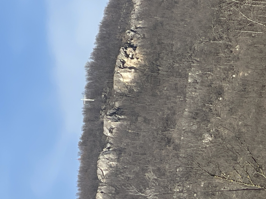 The image displays a panoramic view of a rocky, forested hillside in Cherni Vrah, Bulgaria, under a clear, light blue sky. A large, white Latin cross stands prominently on a rocky outcrop near the summit, positioned right of center. The hillside is densely covered with bare deciduous trees, suggesting a winter or early spring season. Numerous light-colored rock formations and exposed cliffs are visible through the leafless trees, particularly in the mid-section of the slope. In the upper left background, partially obscured by the tree line, a slender, reddish-orange communication tower with a pointed top is discernible. The scene is illuminated by natural daylight, highlighting portions of the rock faces. No human activity or individuals are present.