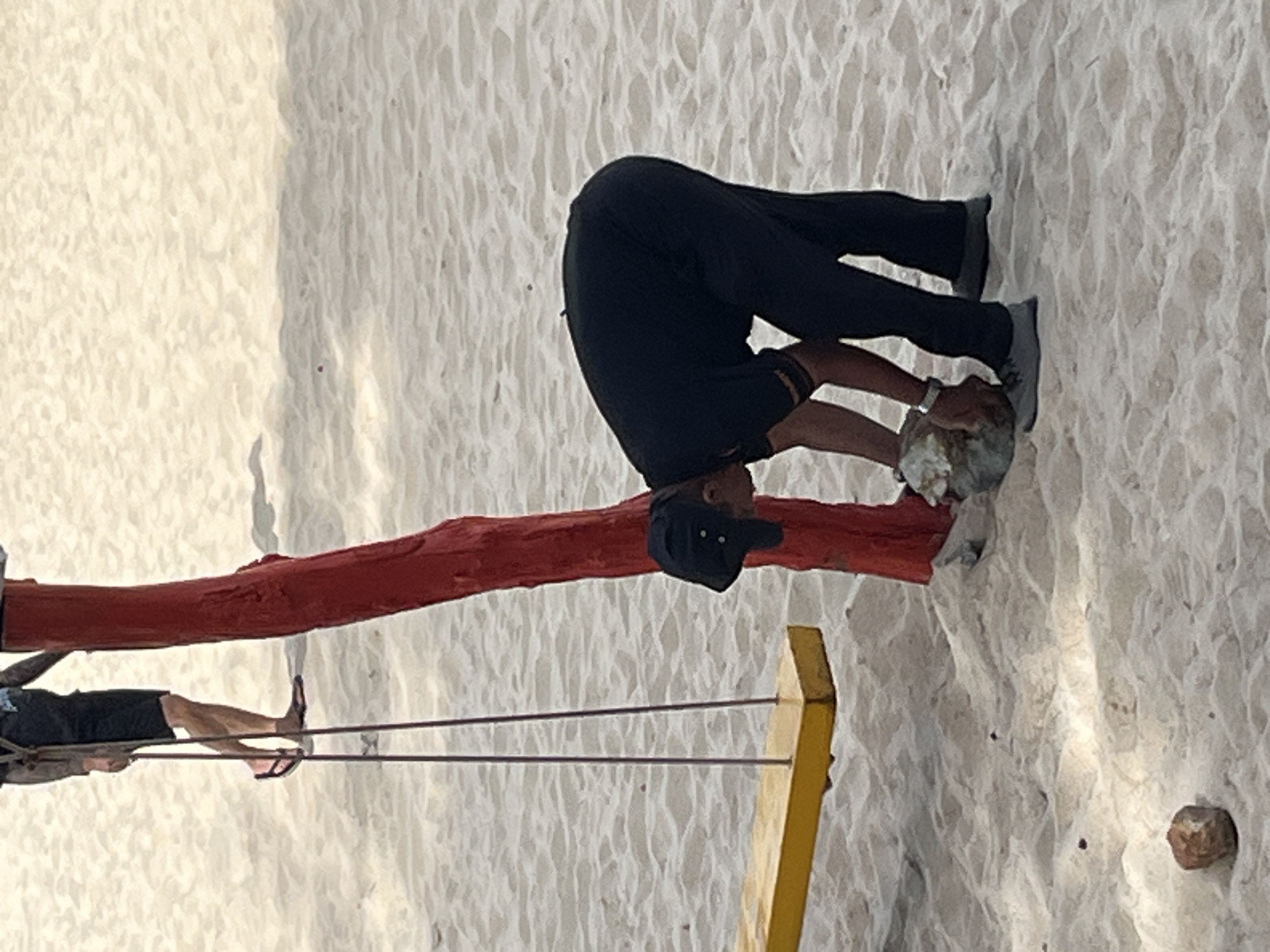 The image depicts a beach scene in Mũi Né, Vietnam. In the foreground, a person wearing a black shirt, black pants, a black cap, and grey shoes is bending down to pick up a rock. The person is also wearing a watch on their left wrist. Beside them, a red post, likely part of a swing set, is visible. A yellow swing seat hangs from the post by two purple ropes. In the background, another person wearing dark shorts and flip-flops is partially visible, standing near the swing set. The ground is covered in light-colored sand, with small rocks scattered on the surface. The lighting suggests it is daytime.