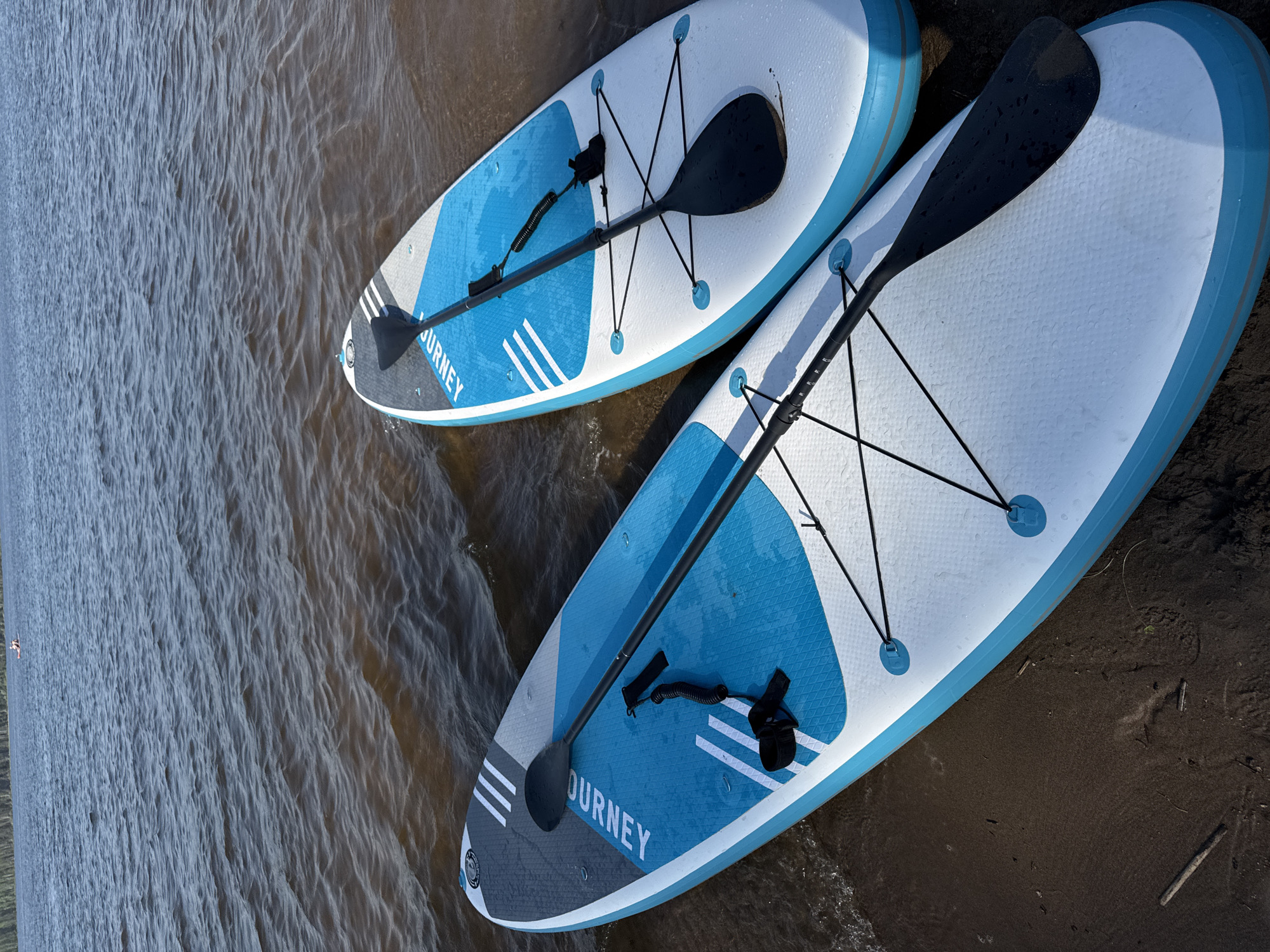 The image shows two inflatable stand-up paddleboards (SUPs) with paddles on the shore of a body of water. The paddleboards are white with blue accents, and each has a paddle resting on top of it, secured by bungee cords. The water is murky brown, and the shoreline is a dark, wet sand or mud. The exact location in Chaikovsky, Russia is not discernible from the image.