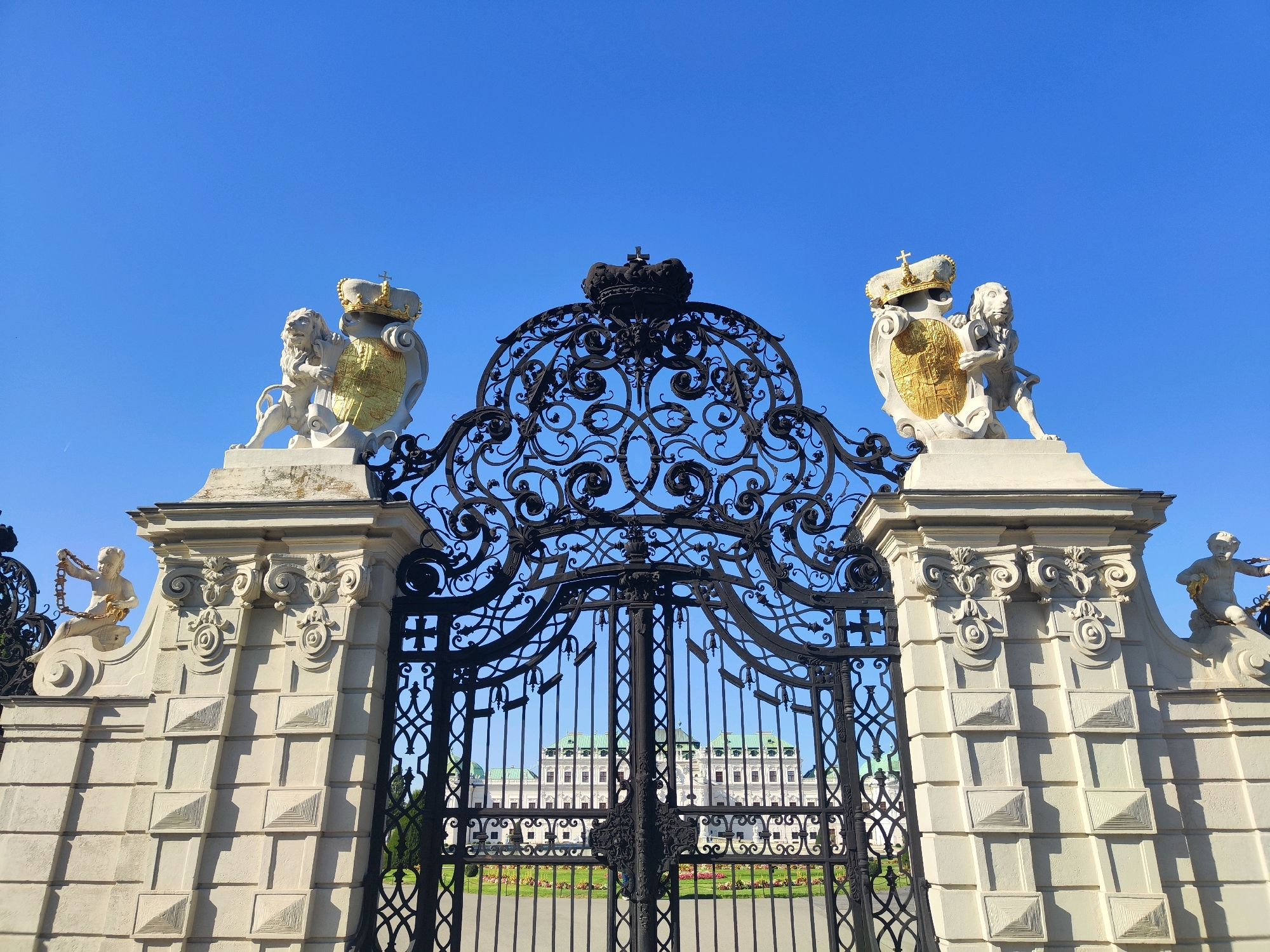 The image shows a grand, ornate black iron gate, likely leading into a park or palace grounds, under a clear blue sky. The gate is flanked by two stone pillars topped with white sculptures of lions holding golden shields with crowns. These pillars are decorated with classical architectural elements, including volutes and geometric patterns. A detailed black iron crown sits at the apex of the gate. Through the gate, a large, white building with a green roof and a dome is visible, surrounded by manicured gardens. This scene is identifiable as the entrance to Schönbrunn Palace in Vienna, Austria.