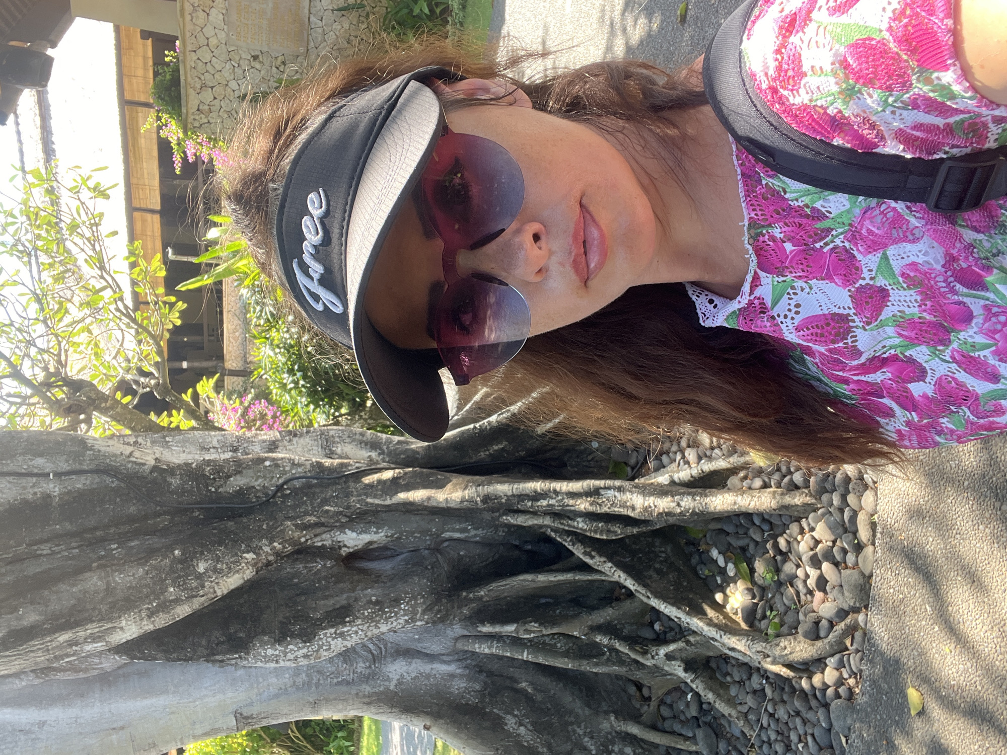 A woman with long, auburn hair is taking a selfie outdoors. She is wearing a black visor with the word "Free" embroidered in white, pink-tinted sunglasses, and a white top with a pink floral pattern. A black backpack is visible over her right shoulder. She is standing next to a large tree with exposed roots, which are covered in small, dark stones. In the background, there is lush greenery, a stone wall, and a building with a thatched roof. The scene appears to be a resort or a garden in Kuta Selatan, Indonesia. The lighting suggests it is a sunny day.