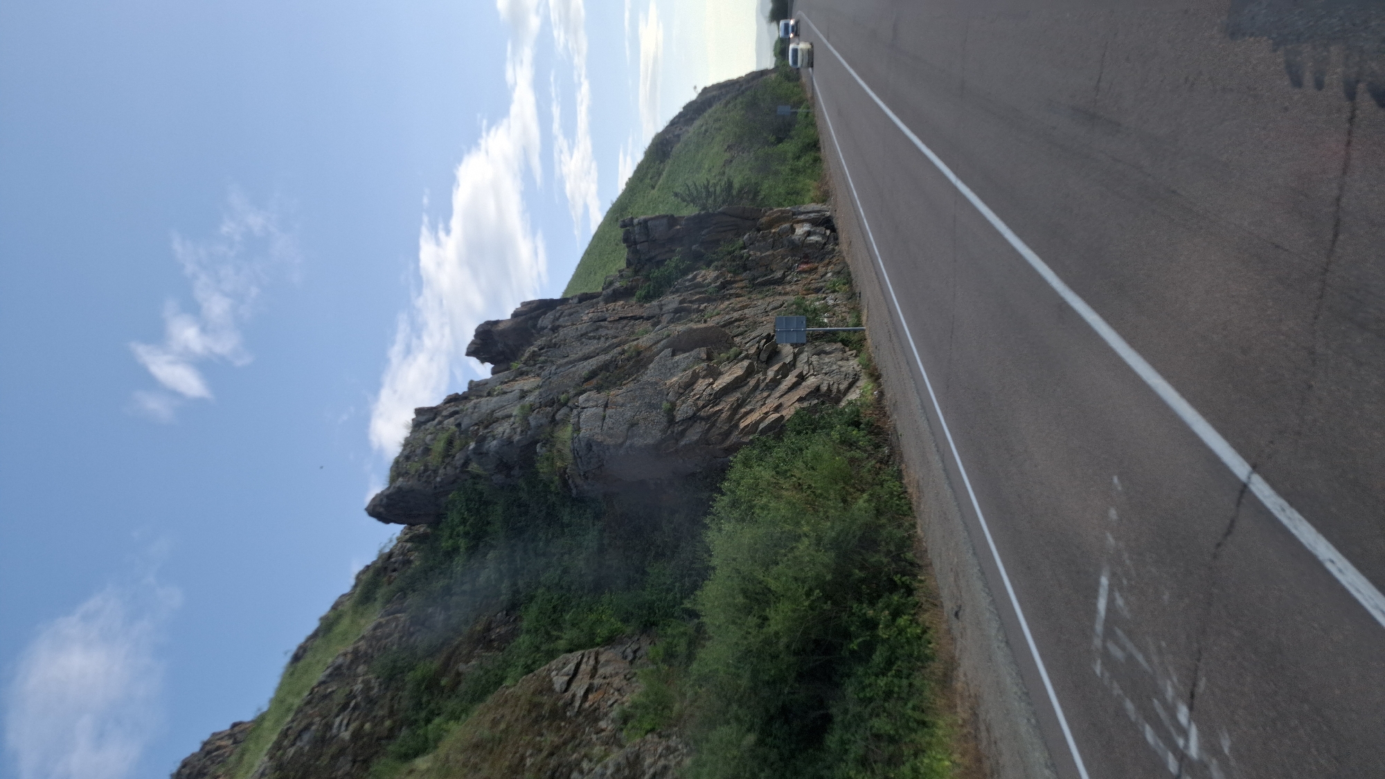 The image shows a scenic view of a road passing by a rocky mountain on a sunny day. The road is paved with asphalt and has white lane markings. On the left side of the road, a steep, rocky mountain covered with green vegetation rises sharply. The mountain has a rugged texture with visible rock formations and some scattered bushes and trees. On the right side of the road, the terrain slopes upwards, also covered in green grass and sparse trees. Two white vehicles are visible driving on the road in the distance. The sky is blue with scattered white clouds. The scene appears to be a natural landscape, possibly a mountain pass or a road through a hilly region. The image was taken in the city of Tarbagatay, Russia.