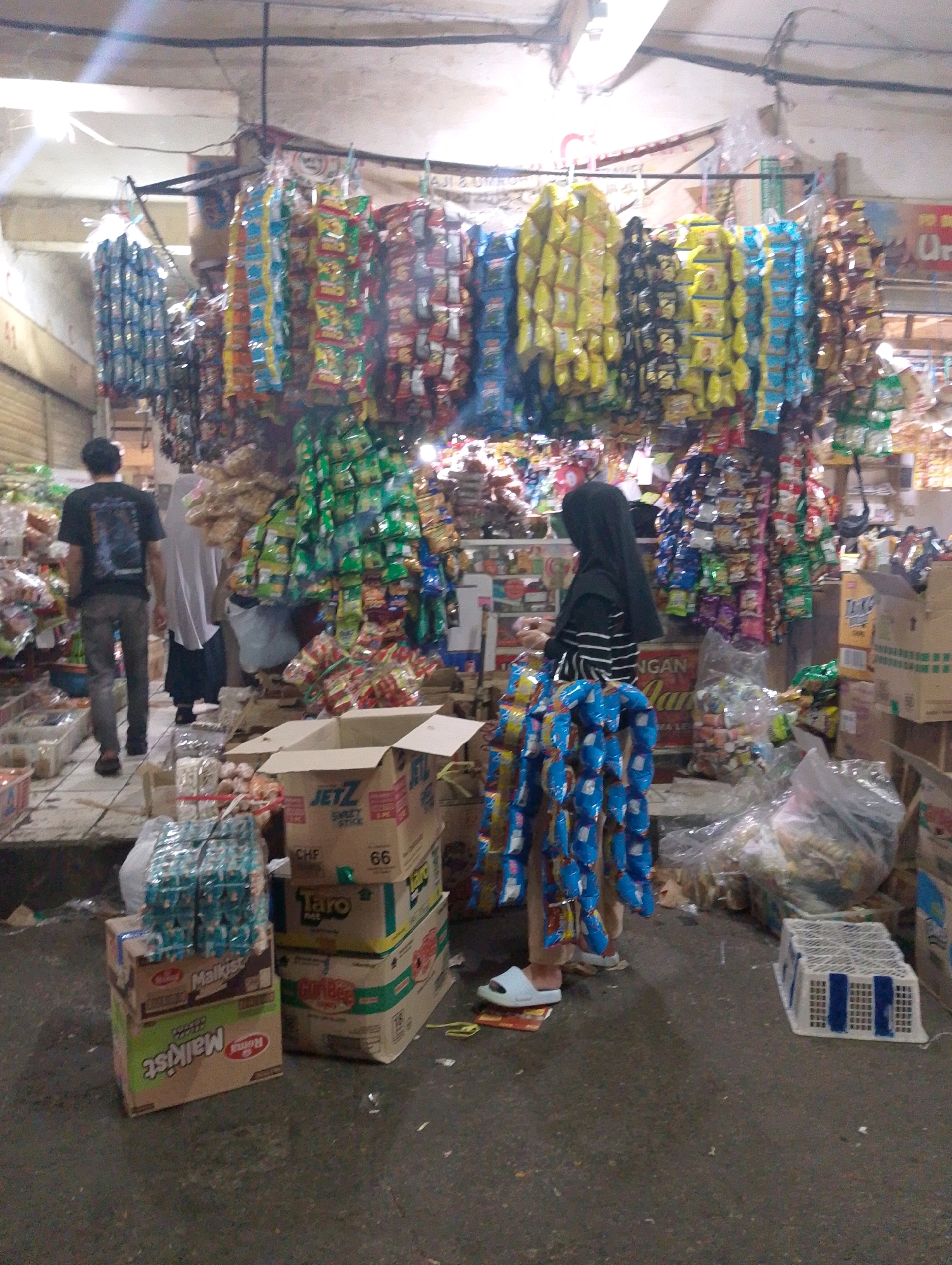 The image depicts an indoor market stall in Lembang, Indonesia, overflowing with various packaged goods, primarily snacks and candies. The stall is densely packed with merchandise, with items hanging from the ceiling and displayed on shelves. In the foreground, several stacks of packaged snacks are visible, along with an open cardboard box. In the mid-ground, a person wearing a black headscarf and a striped shirt is seen standing near a display of hanging snacks. Further back, another person in a black t-shirt and jeans walks away from the camera, and a third person in a white headscarf is also visible. The environment appears to be a busy market, with goods stacked high and hanging in colorful arrays. The lighting suggests an indoor setting, possibly illuminated by artificial lights.