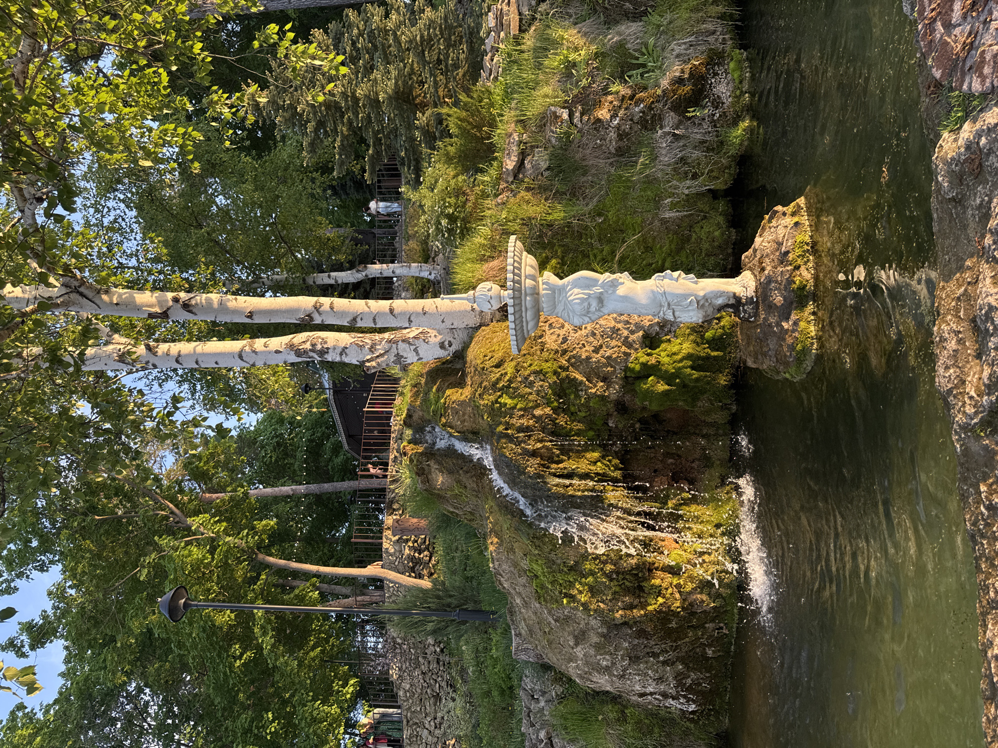 The image depicts a waterfall feature in Krasny Klyuch, Russia. Water cascades over moss-covered rocks into a pool. Atop the rocks, a white statue sits on a tiered platform. Birch trees stand adjacent to the rocks. To the left of the waterfall, there is a stone wall and a lamp post. A set of stairs leading upwards is visible in the background. The environment includes lush greenery and trees surrounding the waterfall.