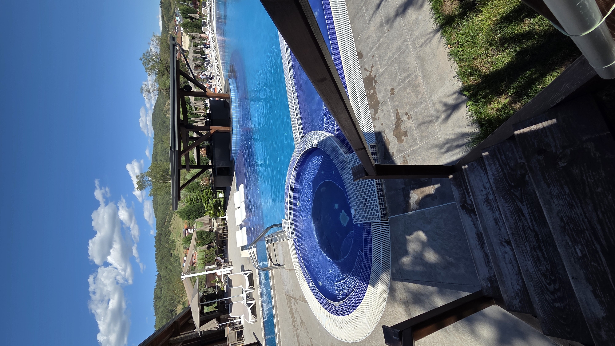 The image shows a swimming pool and hot tub with a blue tiled design. The pool is surrounded by a deck area with gray tiles. Nearby, there is grass and a wooden structure. In the background, there is a mountainous landscape and a clear blue sky with some clouds. There are also some lounge chairs and umbrellas visible near the pool. The location is in the city of Unknown, Unknown.