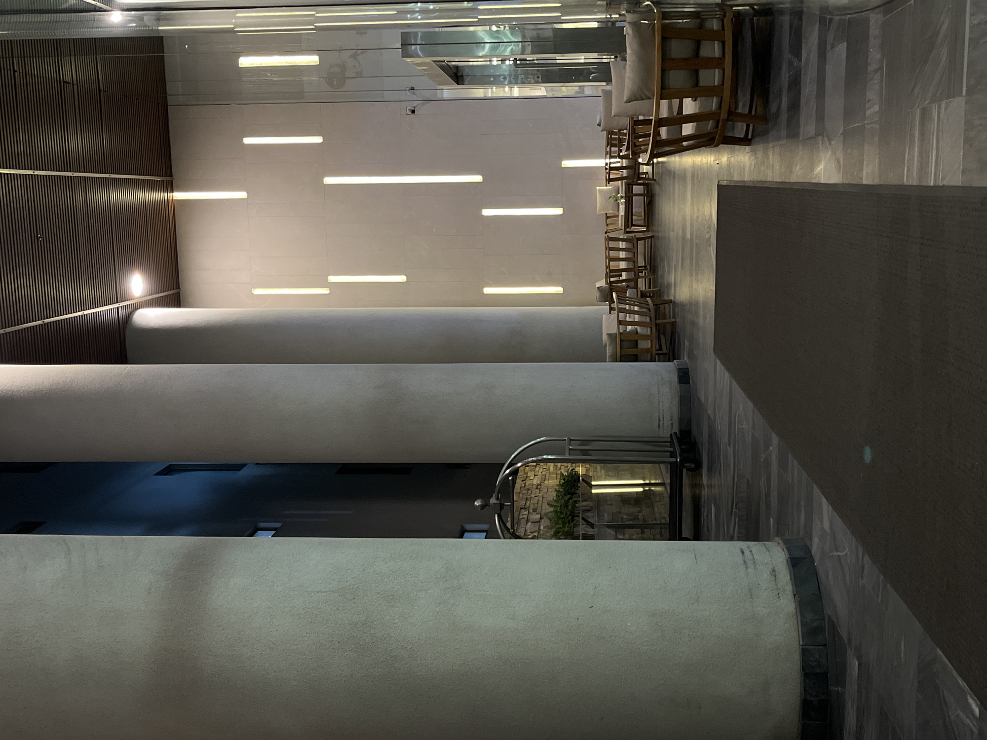 The image depicts the interior of a building, seemingly a hotel lobby, with architectural features including three large cylindrical columns that dominate the foreground. The walls are a combination of dark wood paneling on the left and light-colored tiled surfaces with embedded linear light fixtures on the right. The floor is tiled. In the background, chairs are arranged near a wall, and a luggage cart with a plant is positioned between two of the columns. The scene appears to be within an urban setting in Đà Nẵng, Vietnam, but there are no specific landmarks visible to confirm the exact location. No people are visible in the shot. The environment suggests a modern, possibly upscale, commercial or hospitality space.